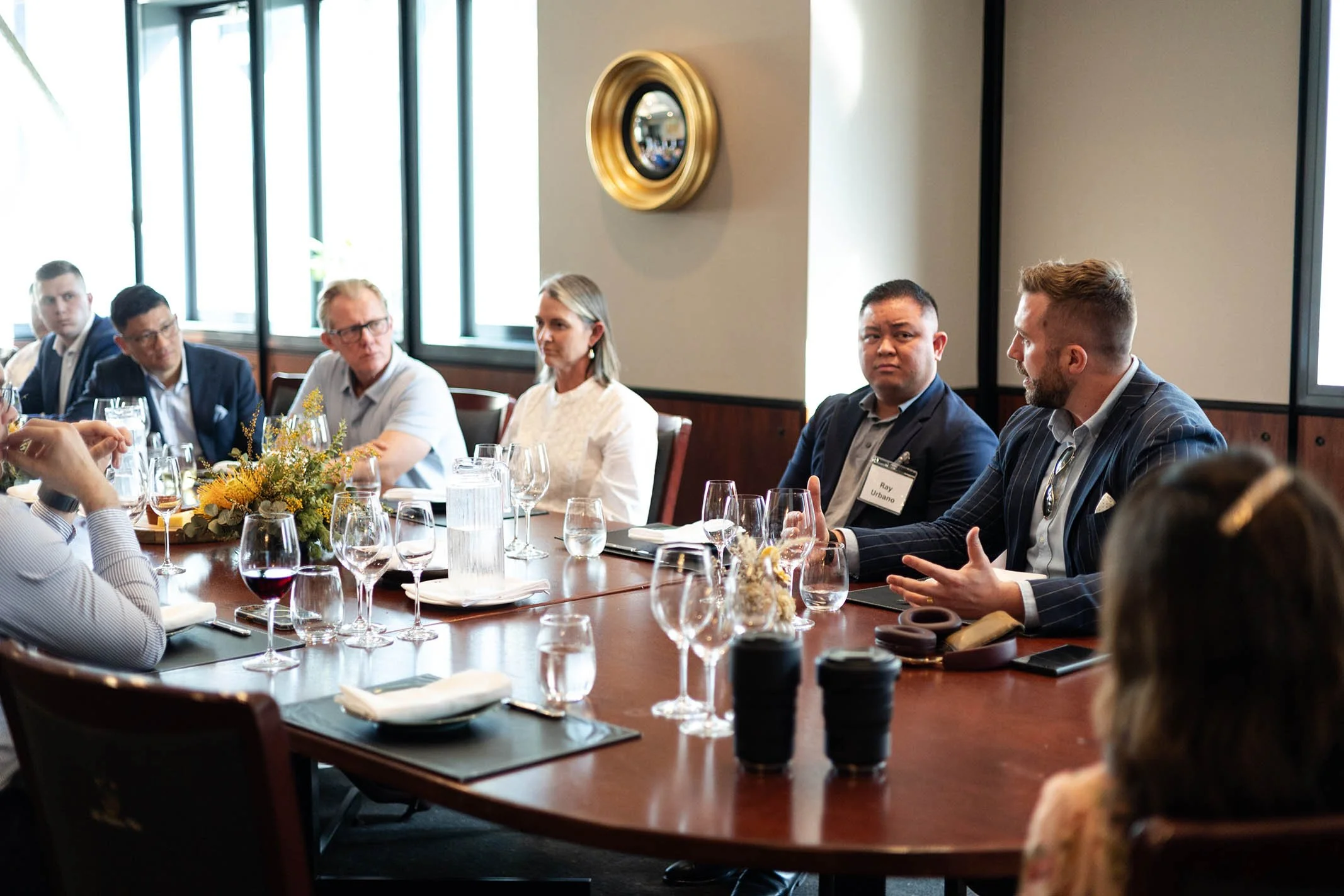 Business professionals seated around a conference table during a meeting, with glasses of water, drinks, and a floral centerpiece.