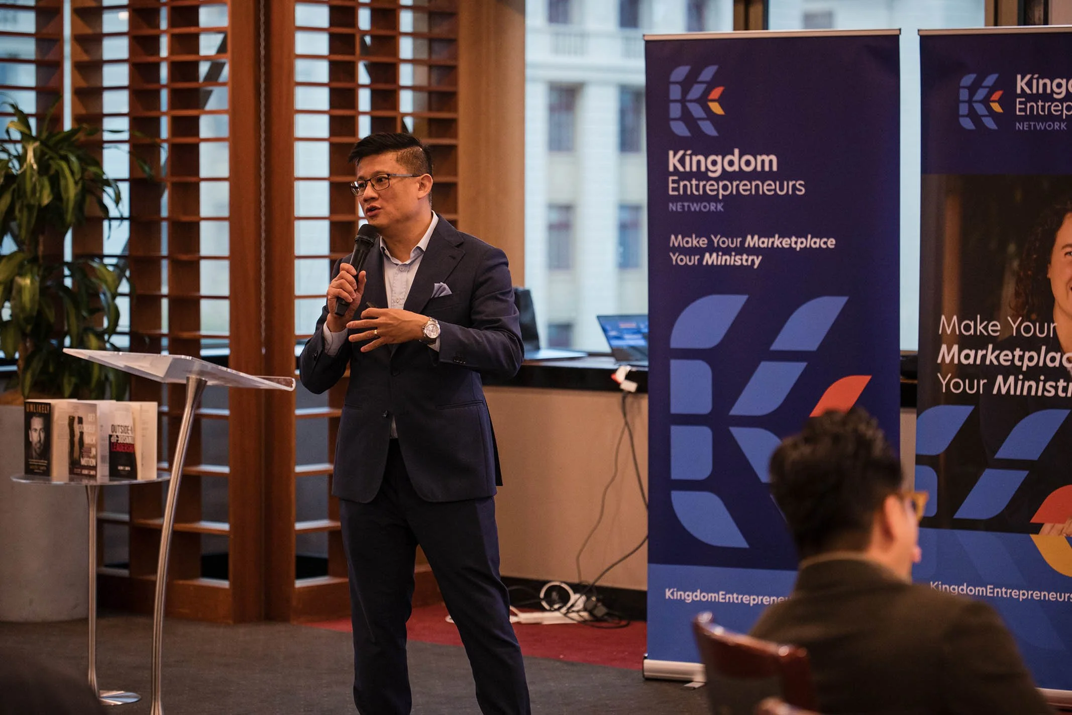 A man in a dark suit with glasses and a watch is speaking into a microphone at a conference. Behind him are banners with the words 'Kingdom Entrepreneurs Network' and the phrase 'Make Your Marketplace Your Ministry.' There are books on a table to his