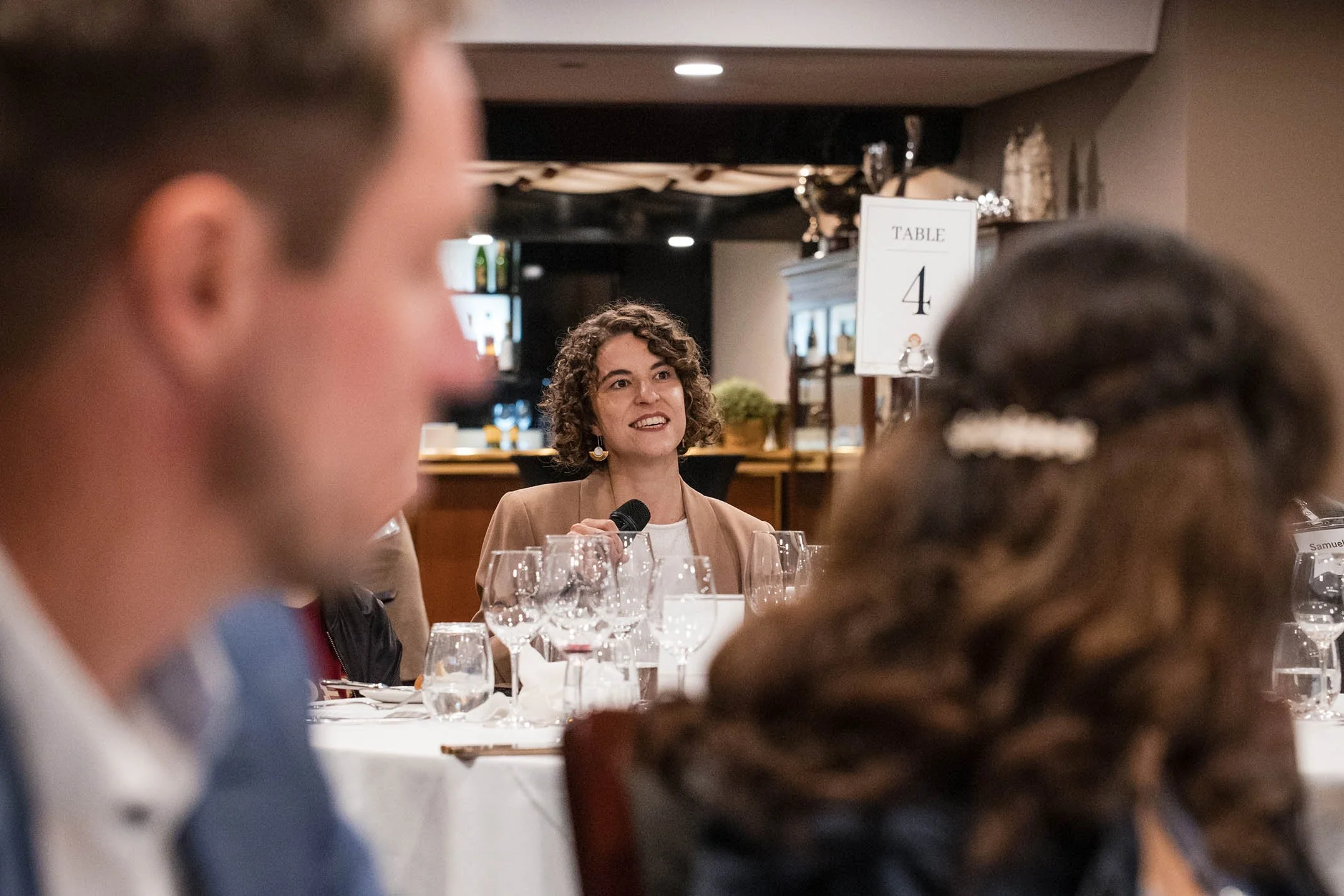 A woman with curly hair and earrings holding a microphone, speaking at a formal event with guests seated at a table with wine glasses, in a restaurant or banquet hall setting.