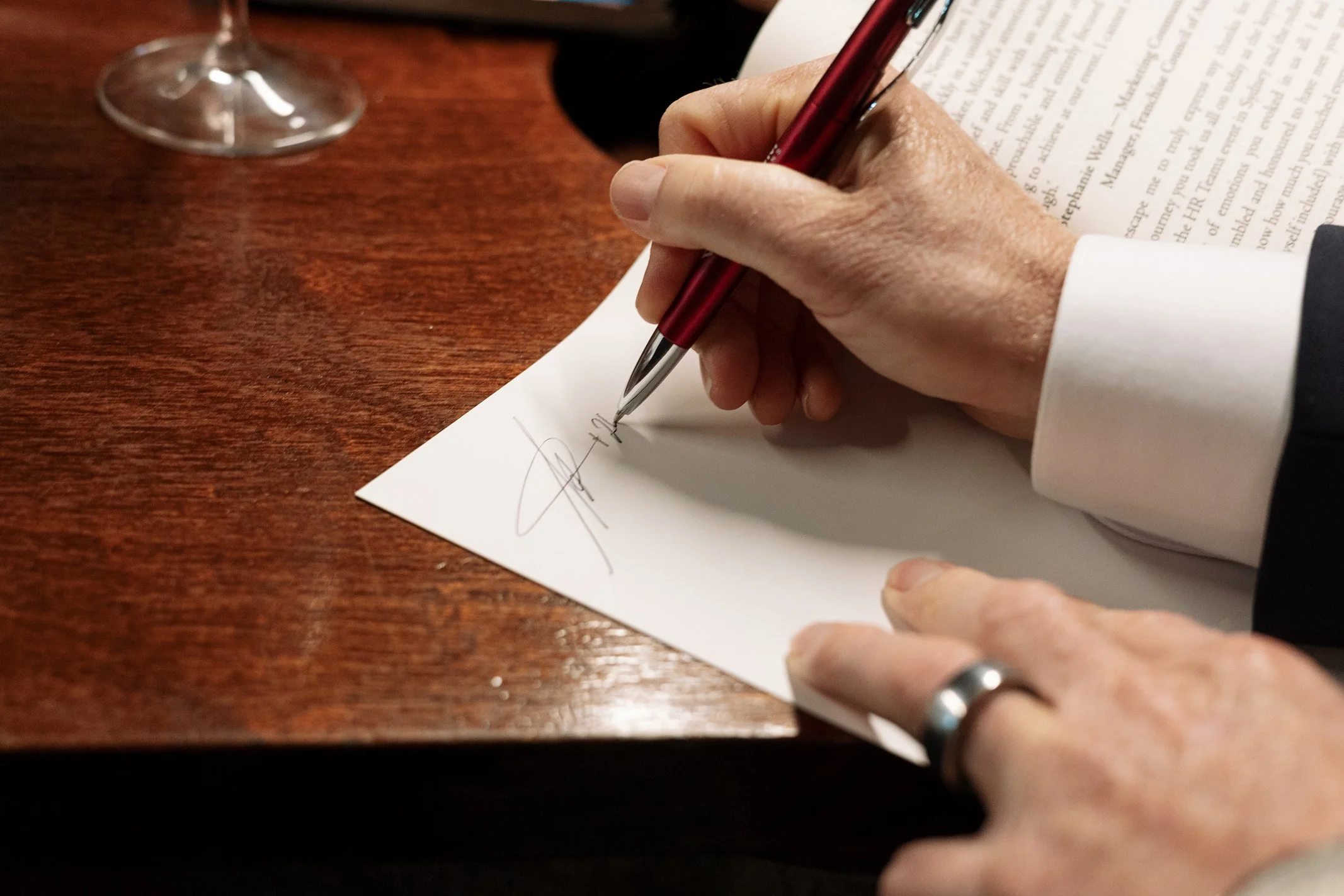 A person signing a document with a red pen on a wooden table, with a glass of wine nearby.