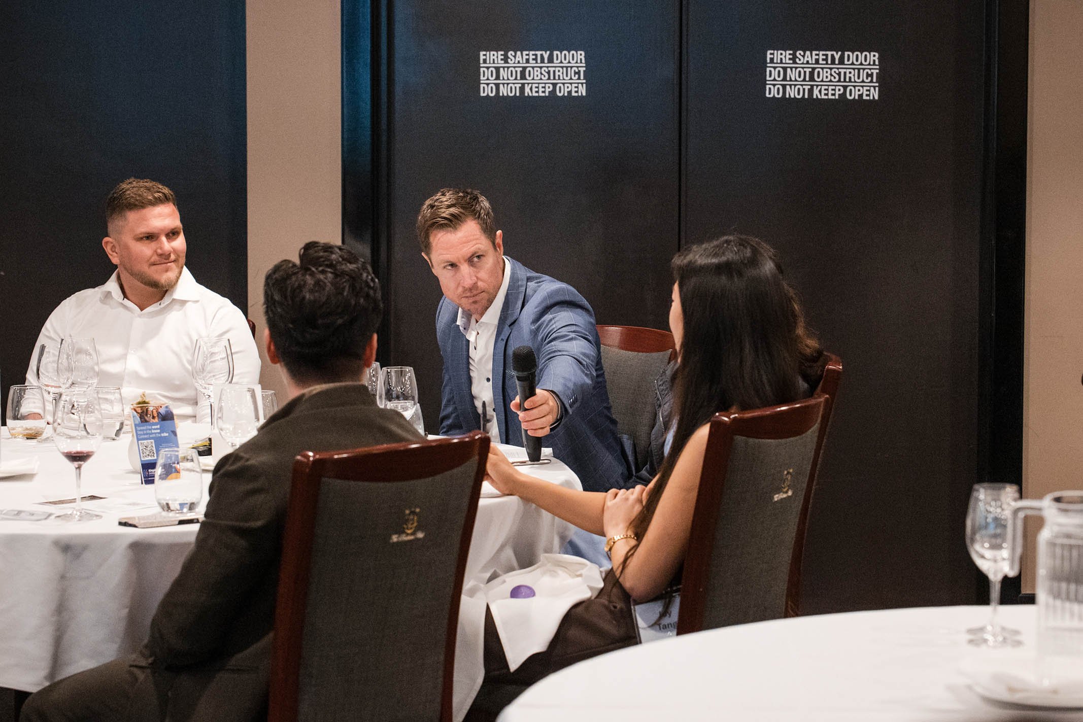 A group of five people sitting around a banquet table, with a man holding a microphone and engaging with a woman in a professional setting.