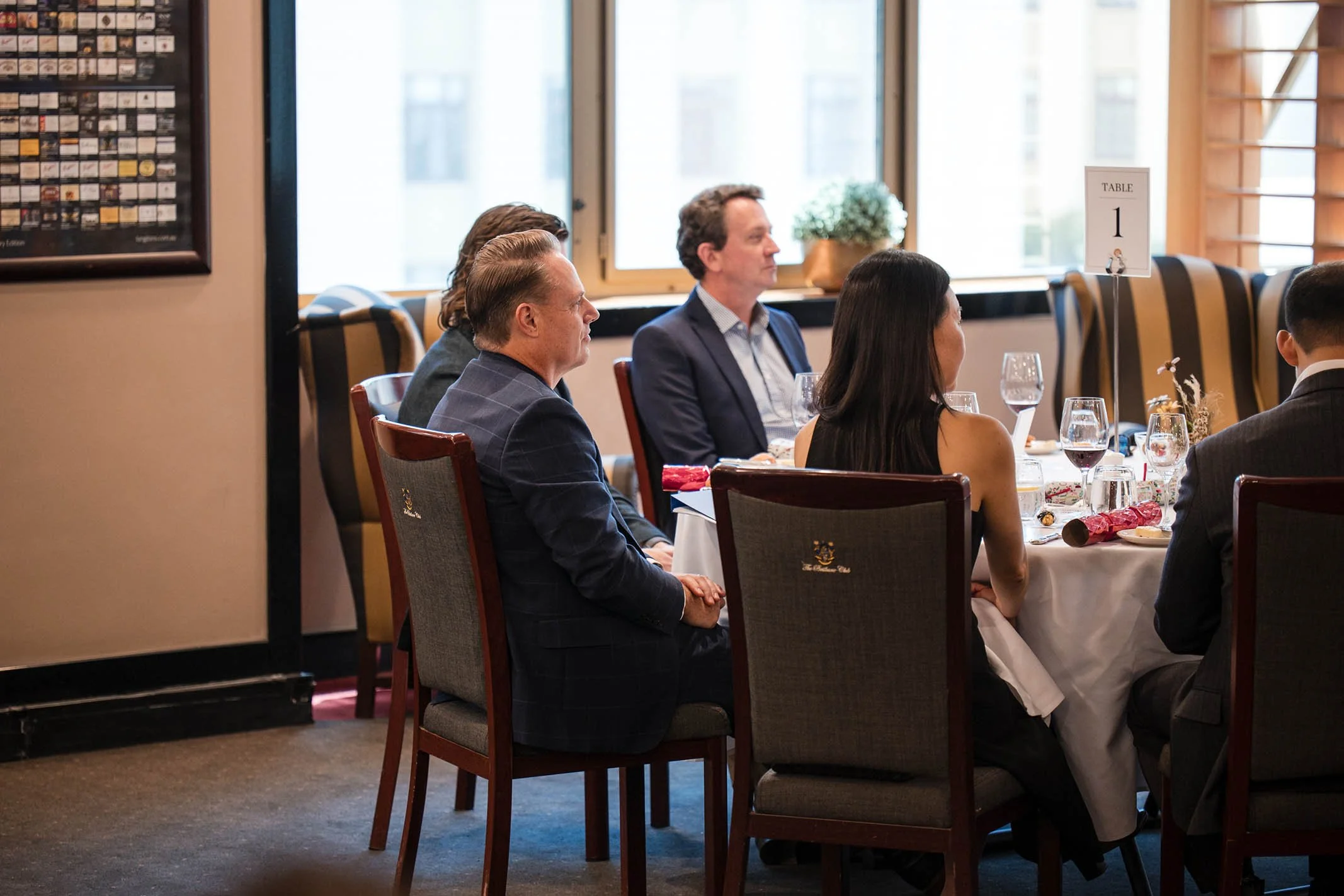 People sitting at a round table decorated for a formal event in a restaurant or banquet hall, with table settings, wine glasses, and a table number sign.