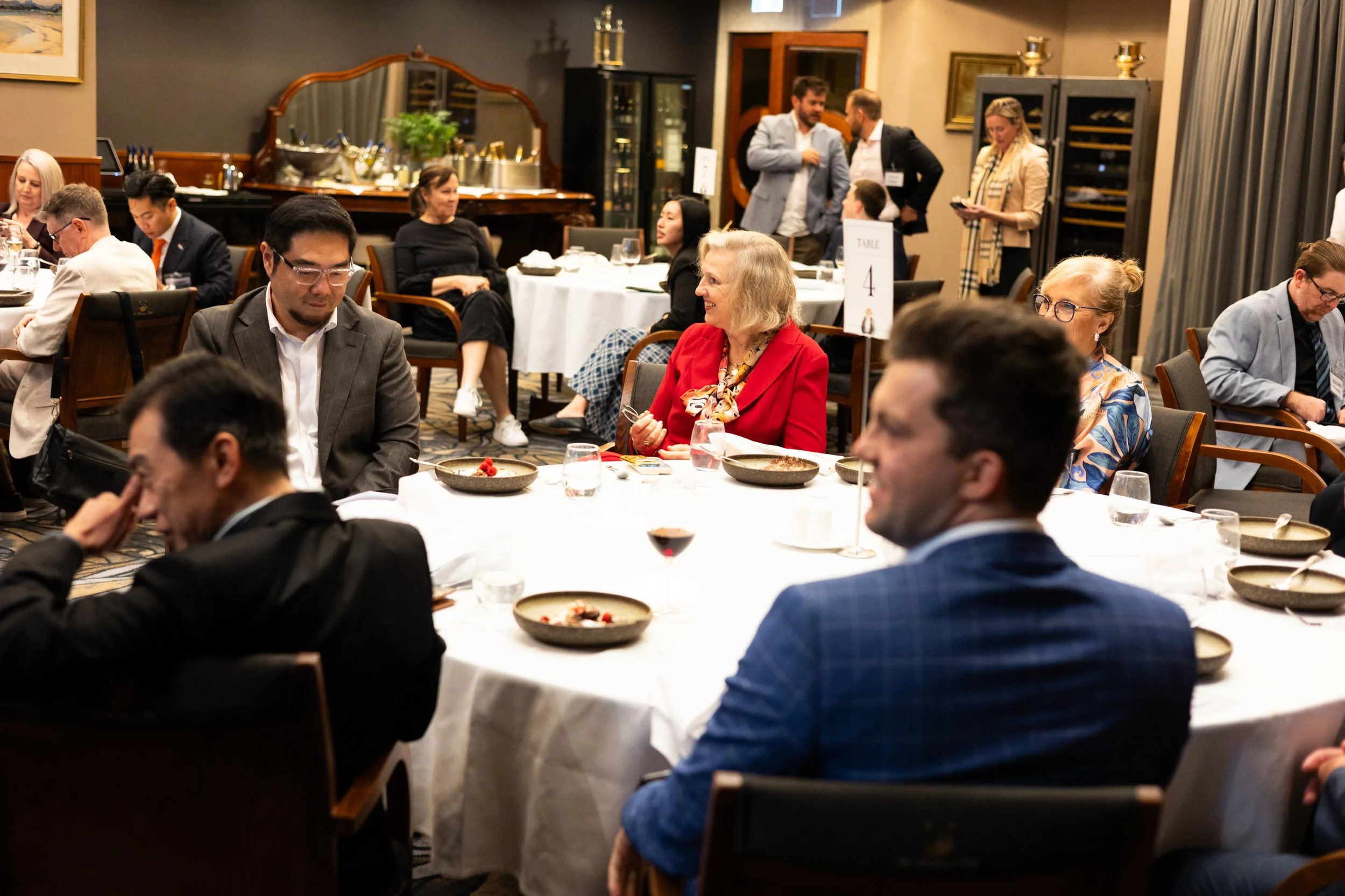 People seated at round tables in a dining room, eating and chatting during a formal event or dinner gathering.