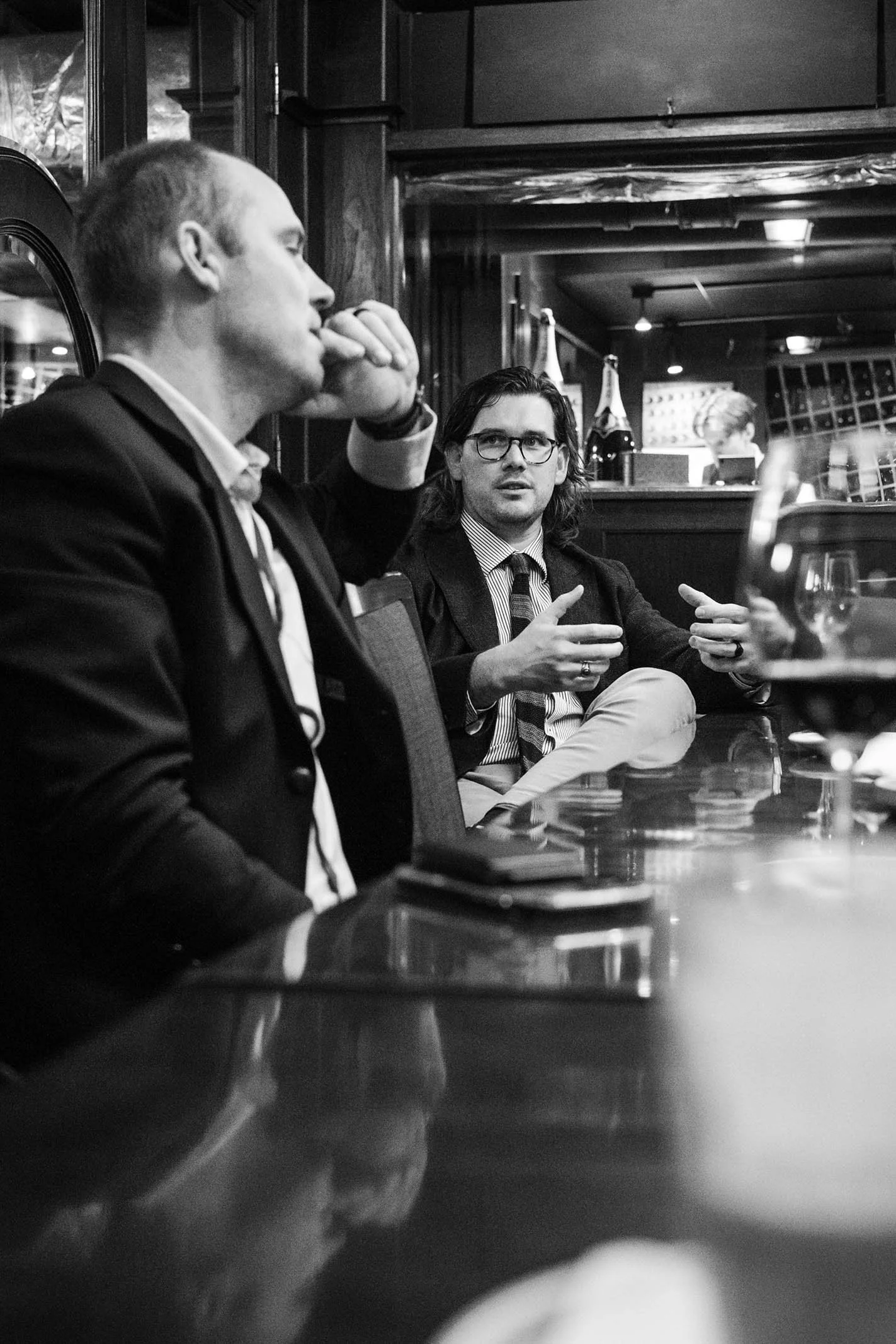 Two men in suits having a conversation in a bar or restaurant. One is listening with his hand on his chin, the other is speaking and gesturing with his hands. There are bottles and glasses in the background.
