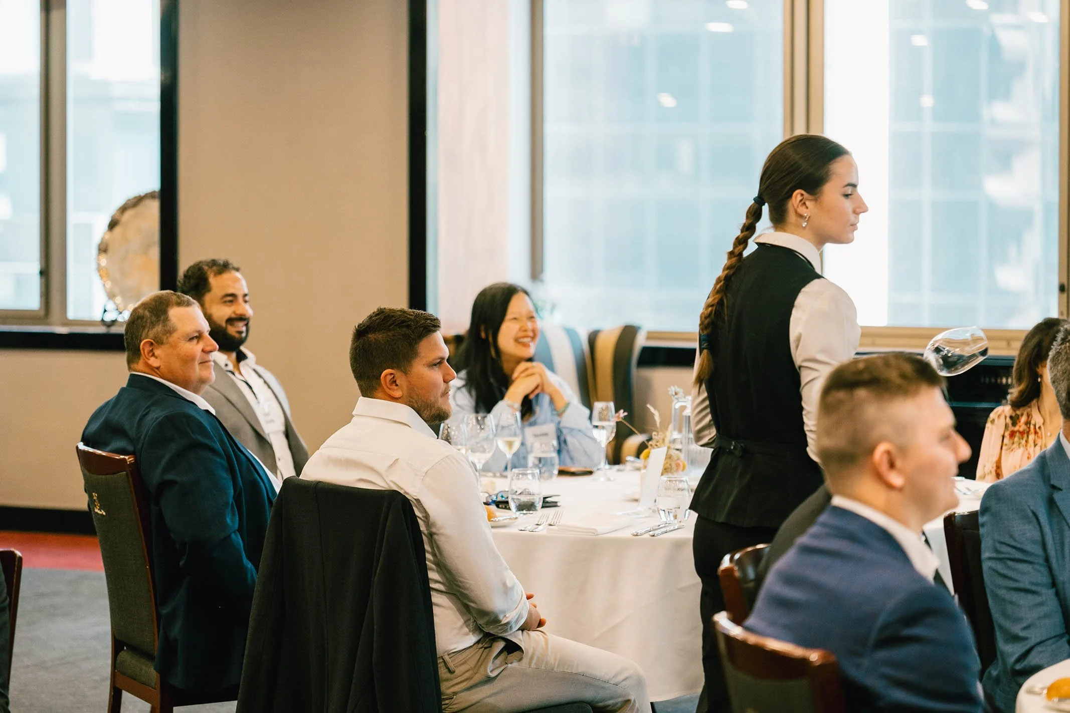 A woman in a black vest and white shirt serving wine at a restaurant table with people clapping and smiling.