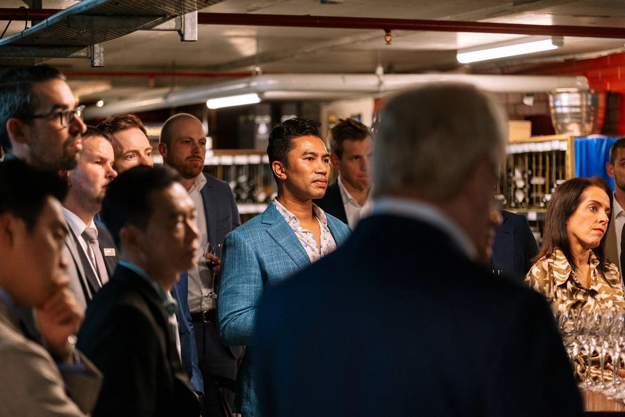 People gathered in a dimly lit warehouse-like space, mostly dressed in formal attire, listening attentively to a speaker or presentation.