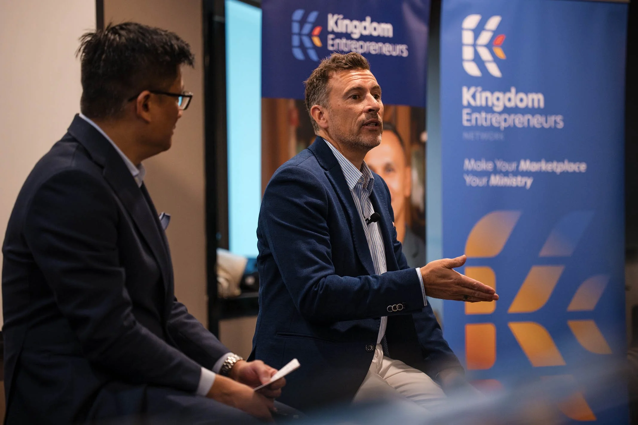 Two men engaged in a discussion at a Kingdom Entrepreneurs networking event. One man is speaking while the other listens, with banners displaying the Kingdom Entrepreneurs logo and tagline in the background.