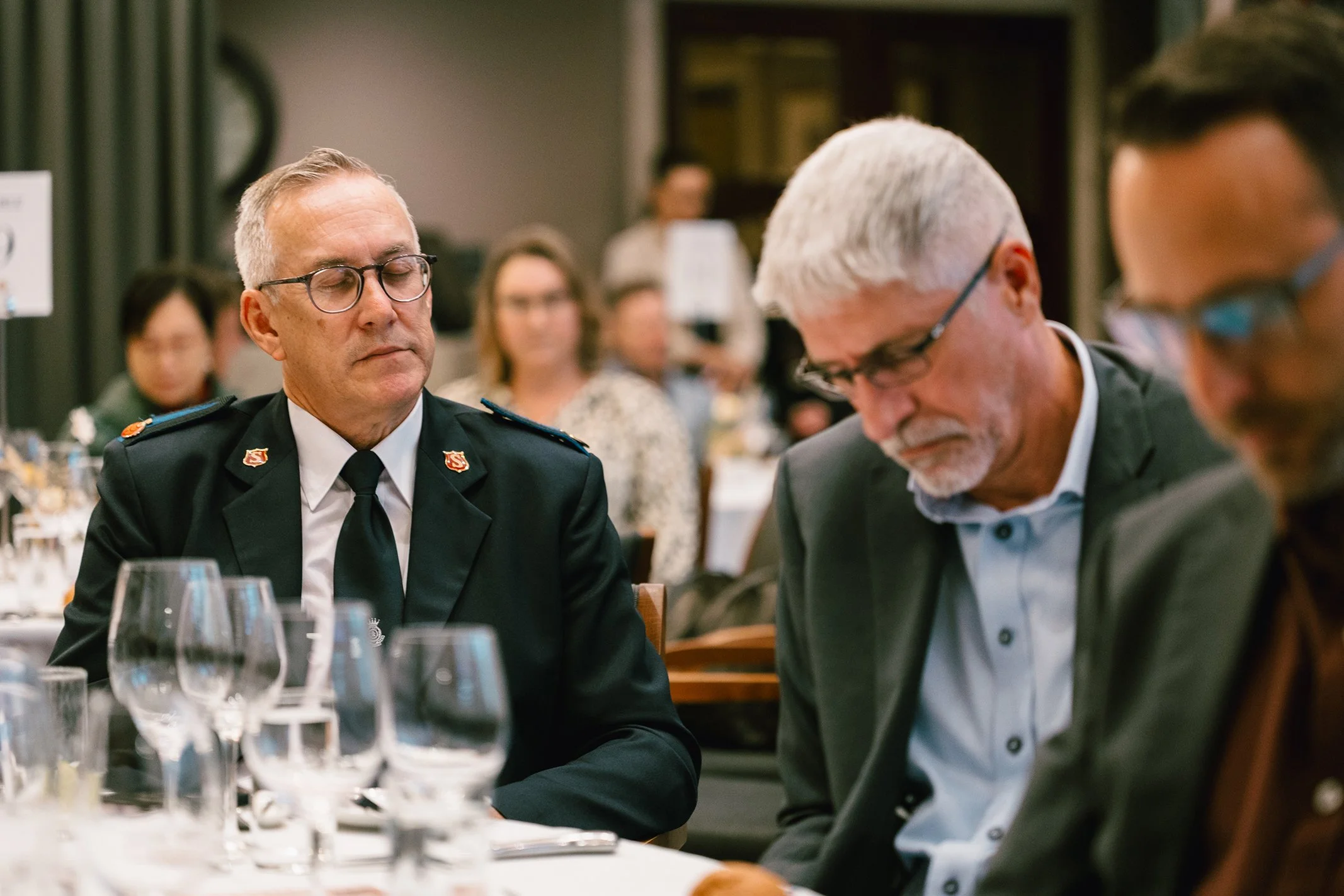 Three men sitting at a table during a formal event, one of them is wearing a sheriff's or military uniform with glasses, the other two are in suits with glasses, surrounded by wine glasses and tableware, with other people in the background.