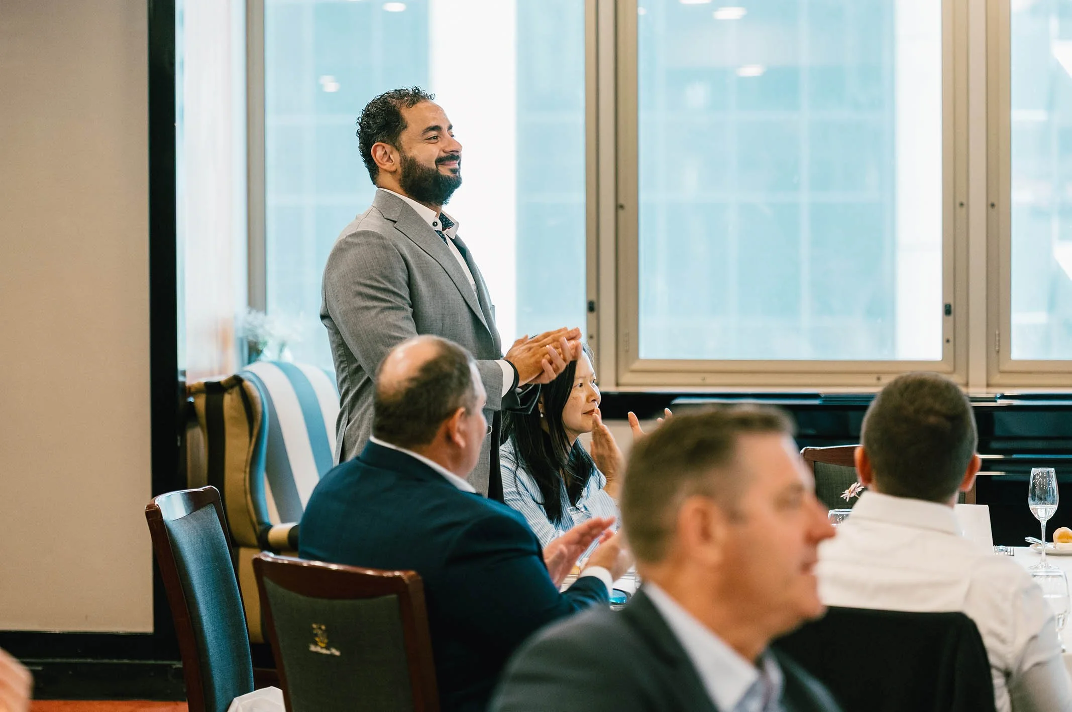 A man in a gray suit standing and smiling during a meeting or conference, with several seated attendees listening, in a room with large windows.