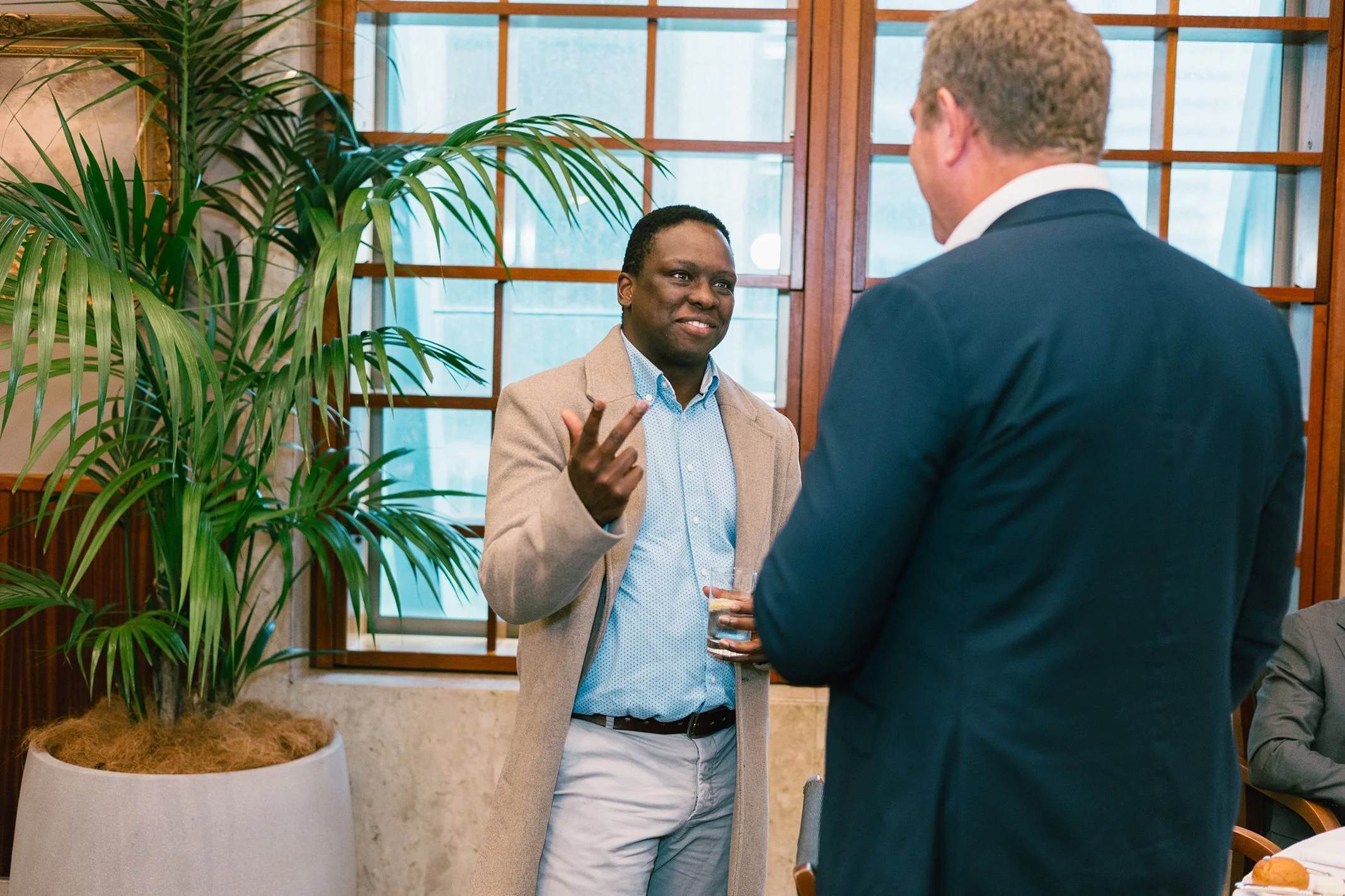 Two men engaged in conversation in a room with large green potted plant, wooden window frames, and natural light.