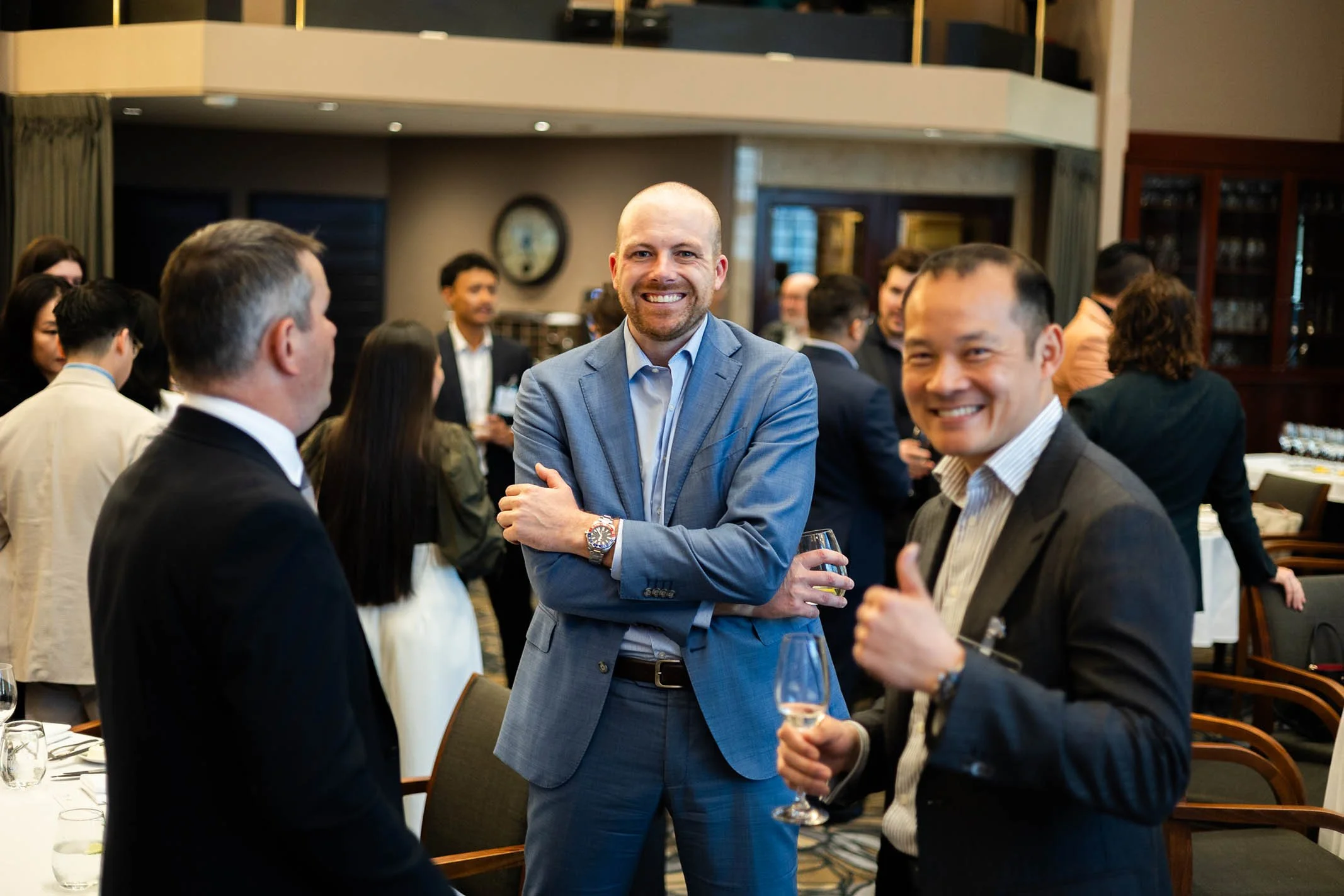 Business professionals mingling at a formal event, some holding glasses of wine, in an elegant venue with wooden furniture and a clock on the wall.