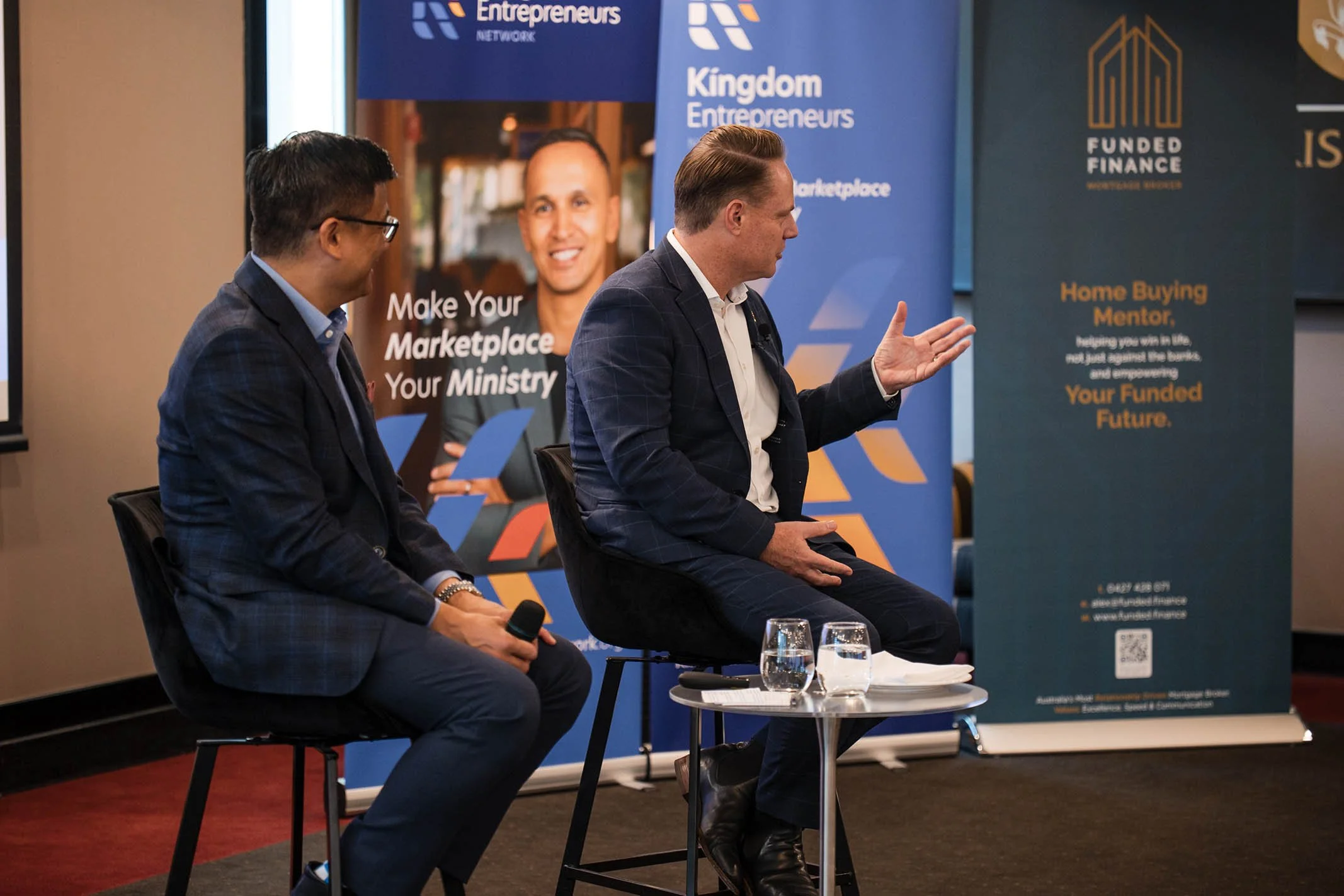 Two men are seated on stage engaging in a discussion at a business event. One is holding a microphone, and behind them are banners with logos and text related to entrepreneurship and finance.