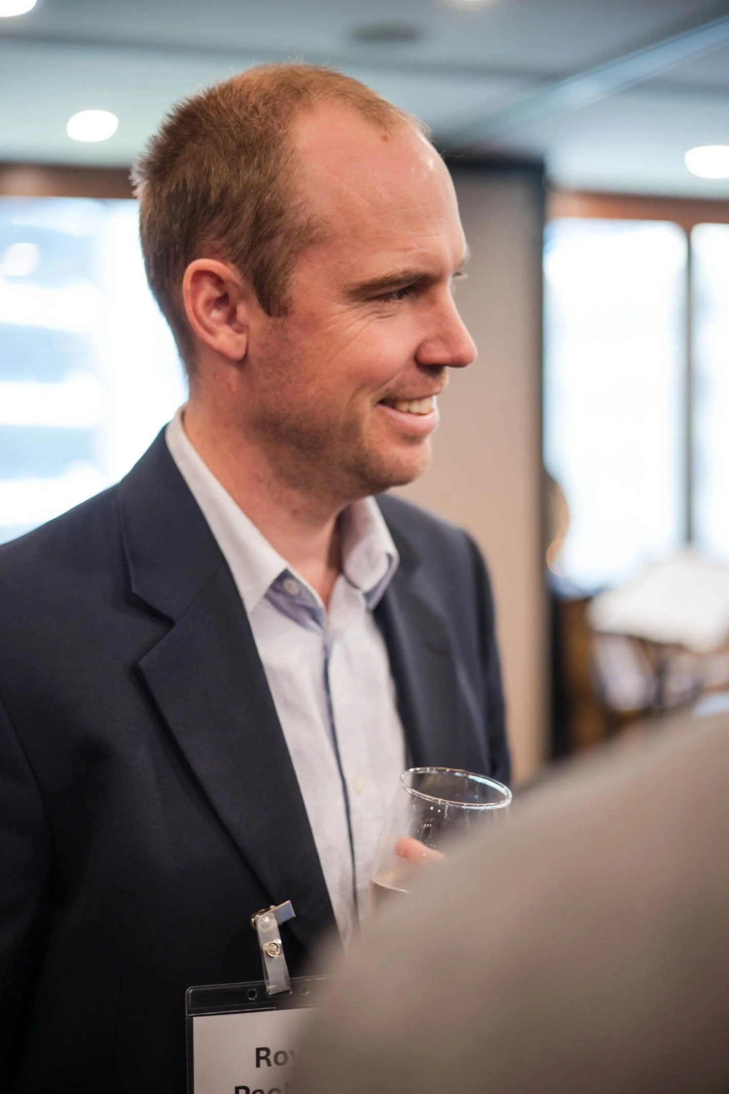 Smiling man in a suit holding a glass of wine at a social or professional event.