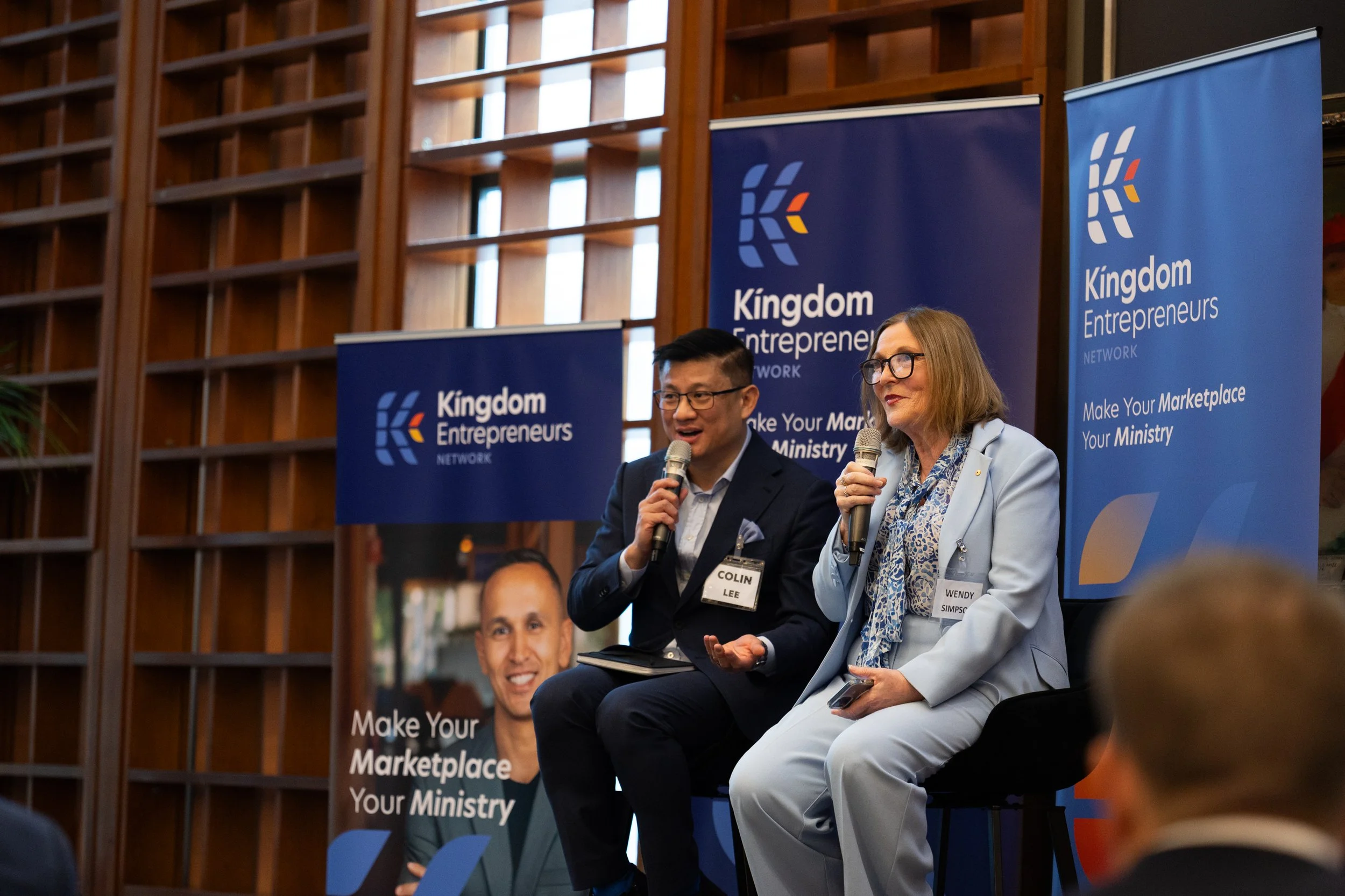 A man and a woman sitting on chairs speaking into microphones during a panel discussion at an event organized by kingdom entrepreneurs, with banners in the background that say "Make Your Marketplace Your Ministry."