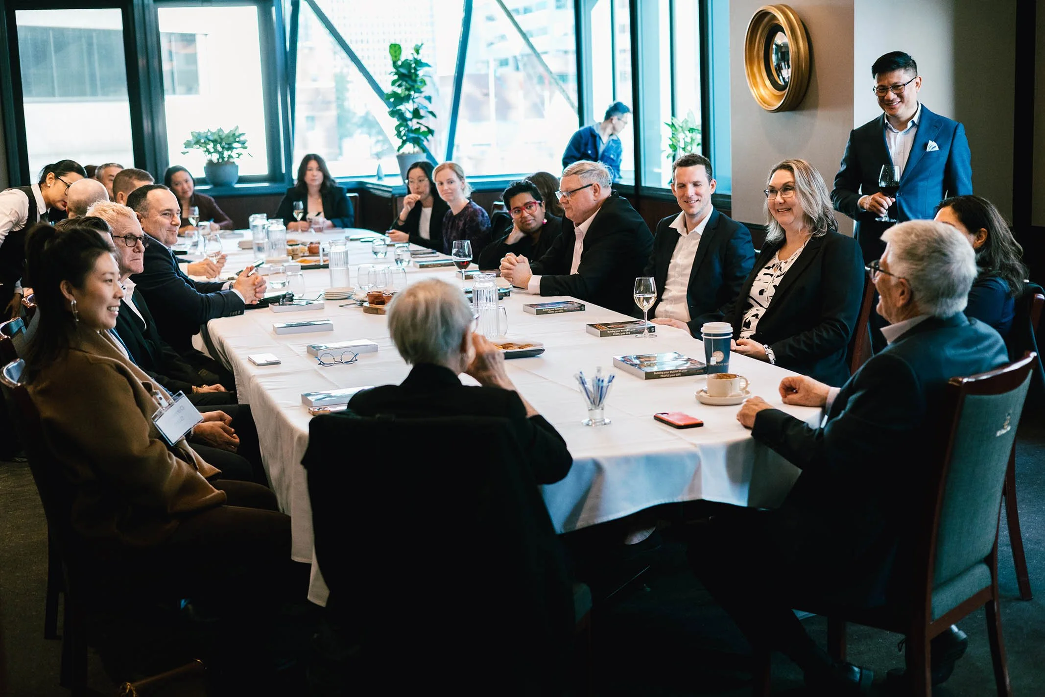 Business meeting with diverse professionals seated around a long conference table in a modern office space, with a man standing and holding a glass of wine.