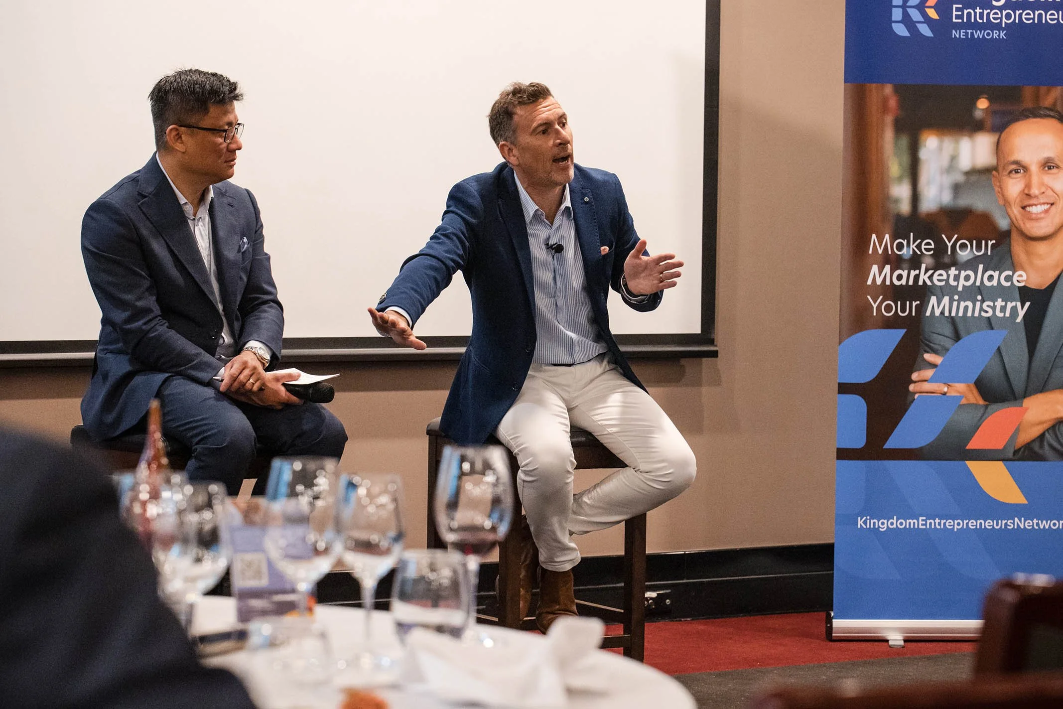 Two men are sitting on stage during a panel discussion. The man on the right is speaking passionately, gesturing with his hands. The man on the left is listening while holding a microphone.
