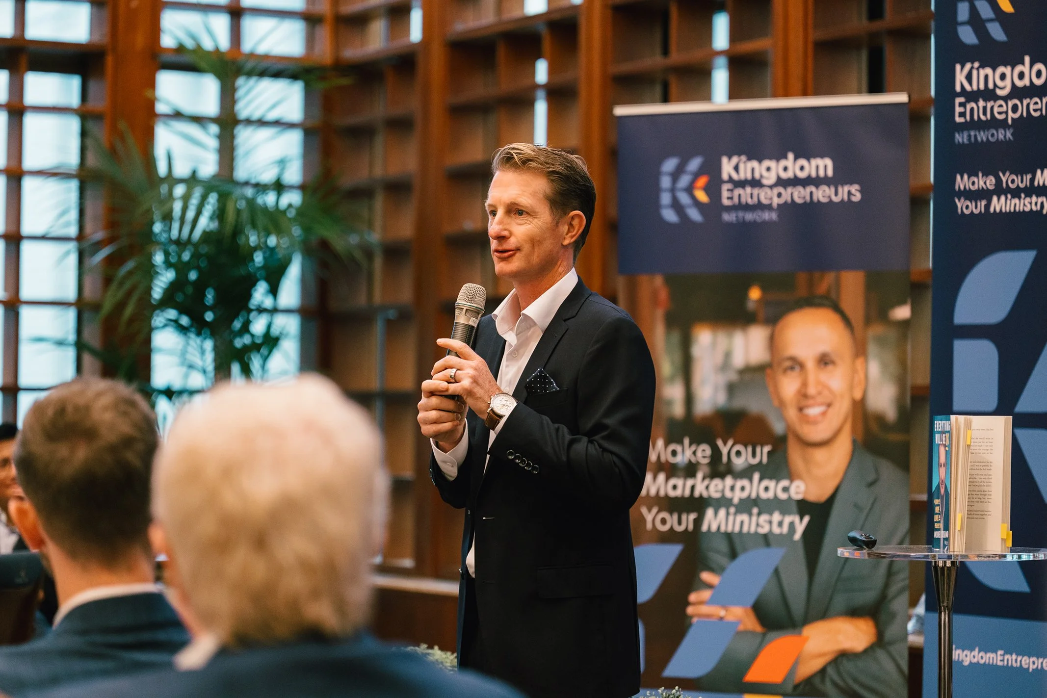 A man in a black suit and white shirt is speaking into a microphone at a conference, with a Kingdom Entrepreneurs banner and crowd in the background.