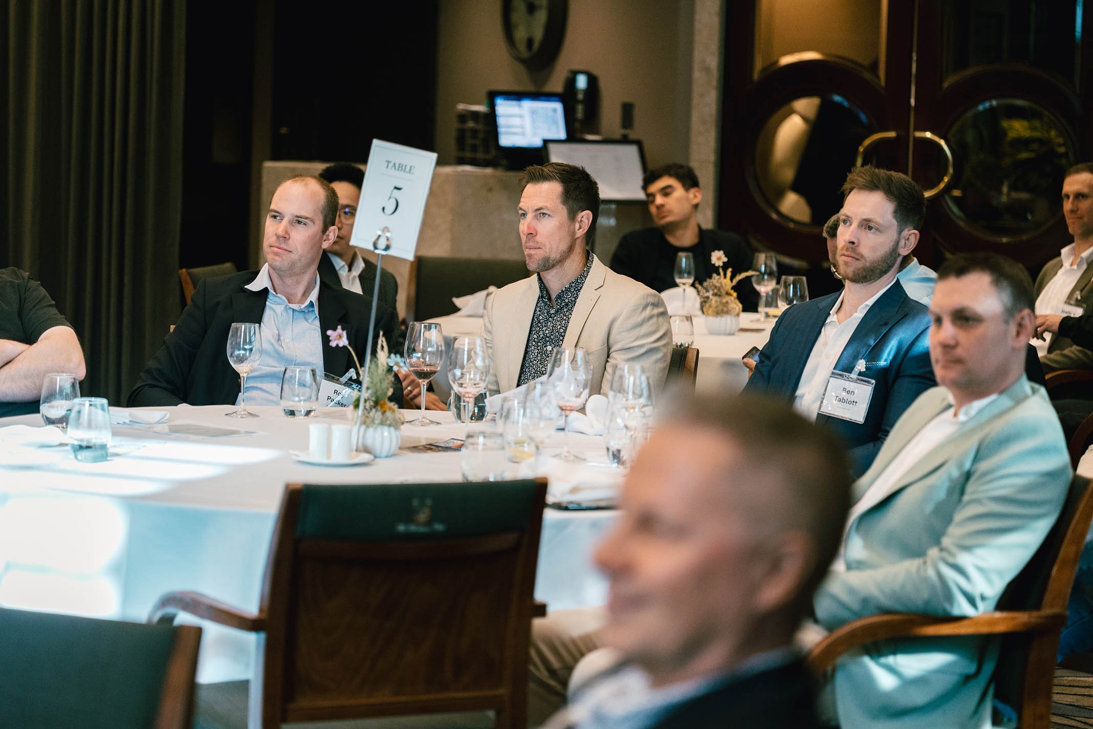 Men attending a formal conference or meeting, sitting at round tables with glasses and floral centerpieces, with a sign indicating table number 5.