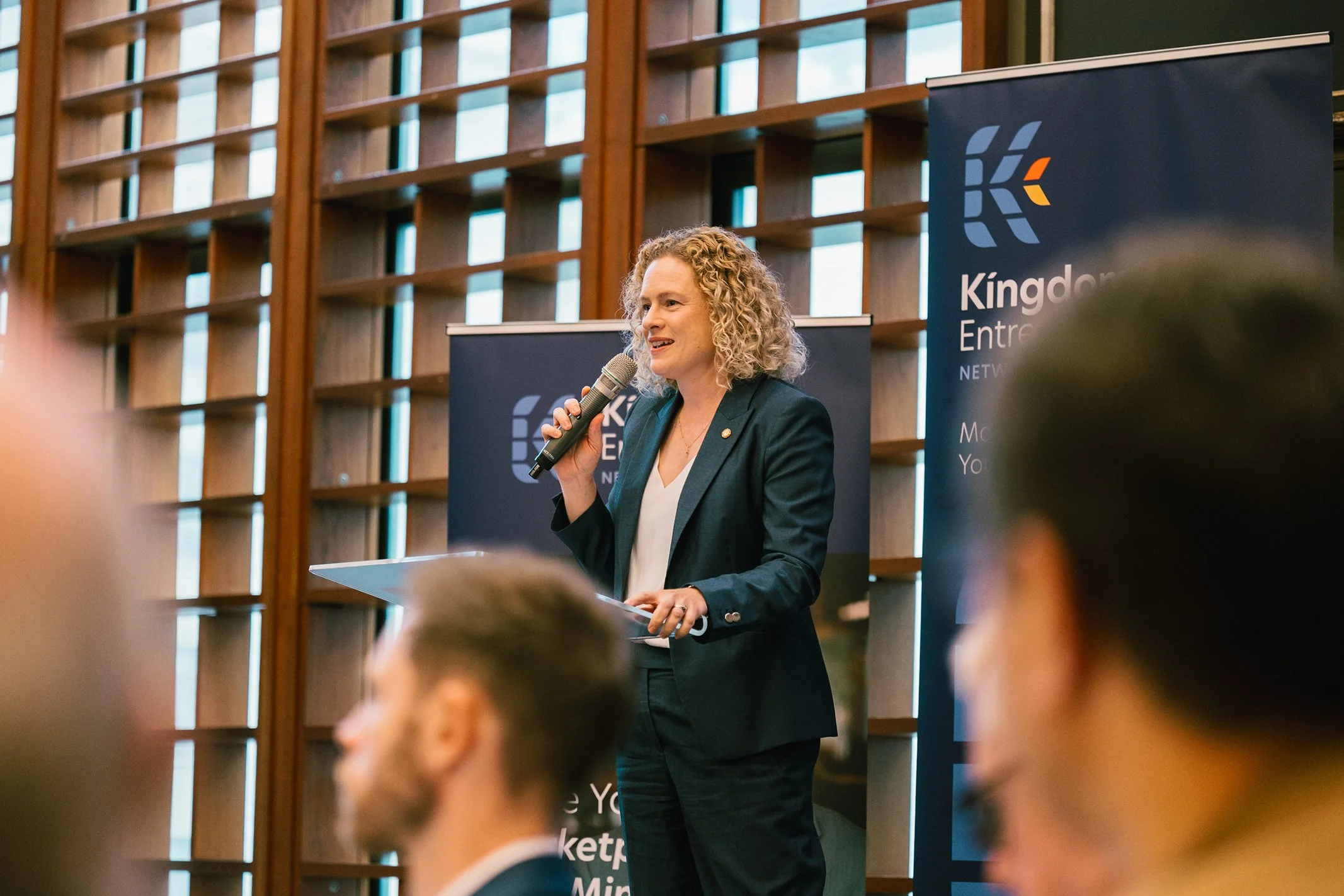 A woman with curly blonde hair in a dark suit speaks into a microphone at a conference. She stands in front of banners that read "Kingdom Entrepreneurship Network" in a room with large wooden-paneled windows.