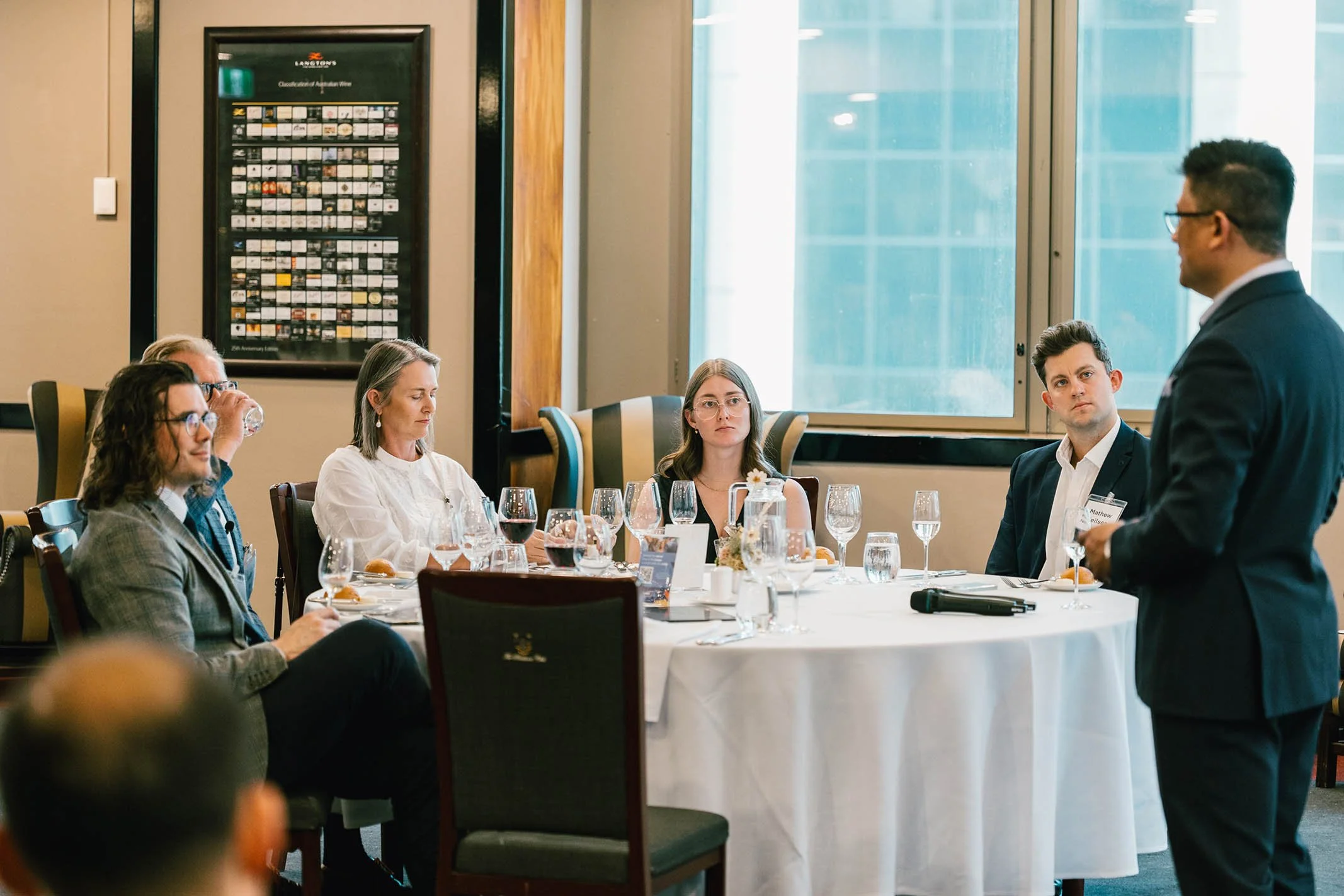 A man in a suit giving a presentation to a group of five people seated around a round table, which is set for a meal with wine glasses and plates, in a conference room with a large window and a framed chart or poster on the wall.