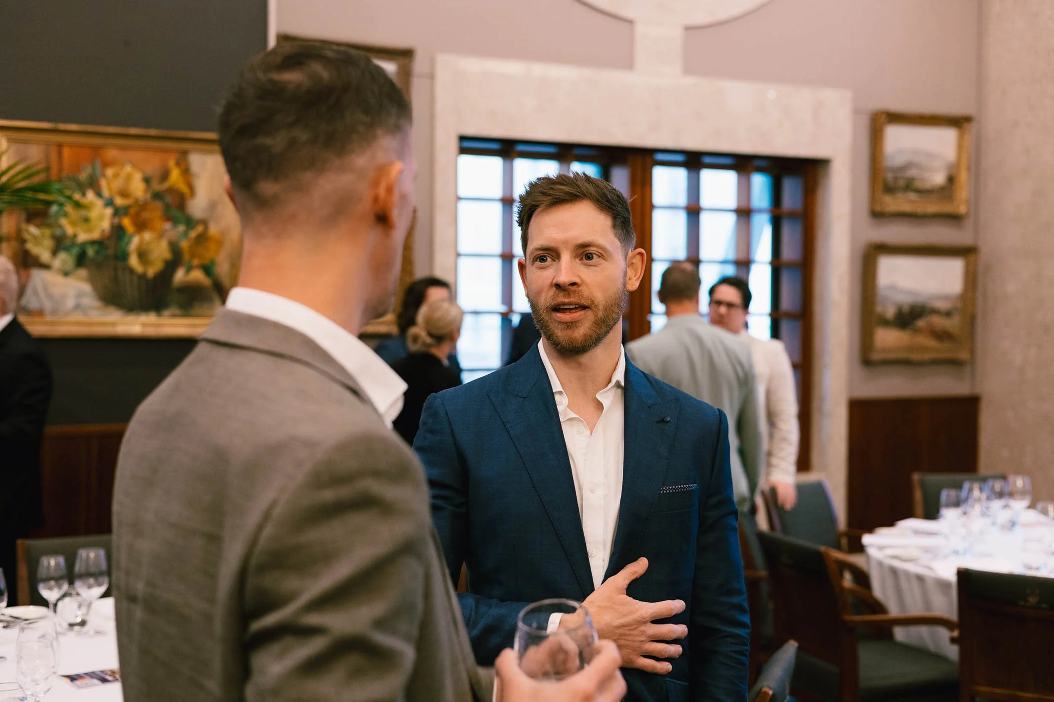 Two men engaged in conversation at a formal event in a decorated dining room with paintings and a window in background.