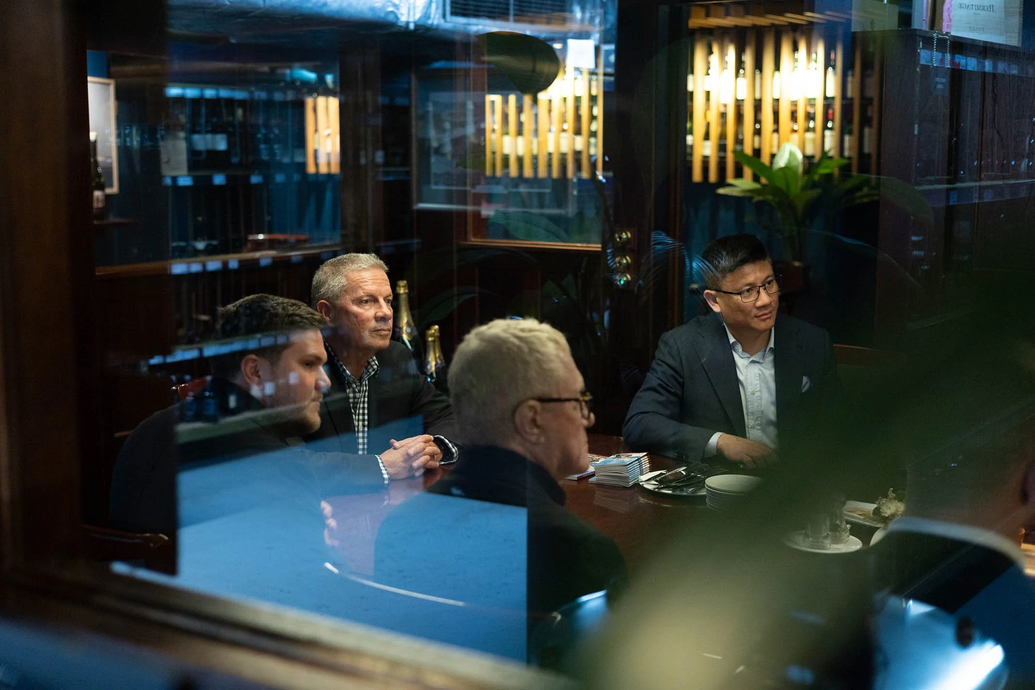People sitting at a table through a glass window in a restaurant or meeting room, engaged in a serious discussion, with bottles and napkins on the table.