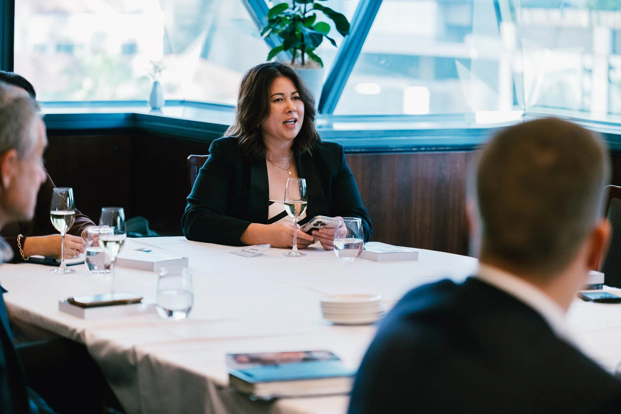 A woman speaking during a business meeting, sitting at a table with glasses of water and a wine glass.