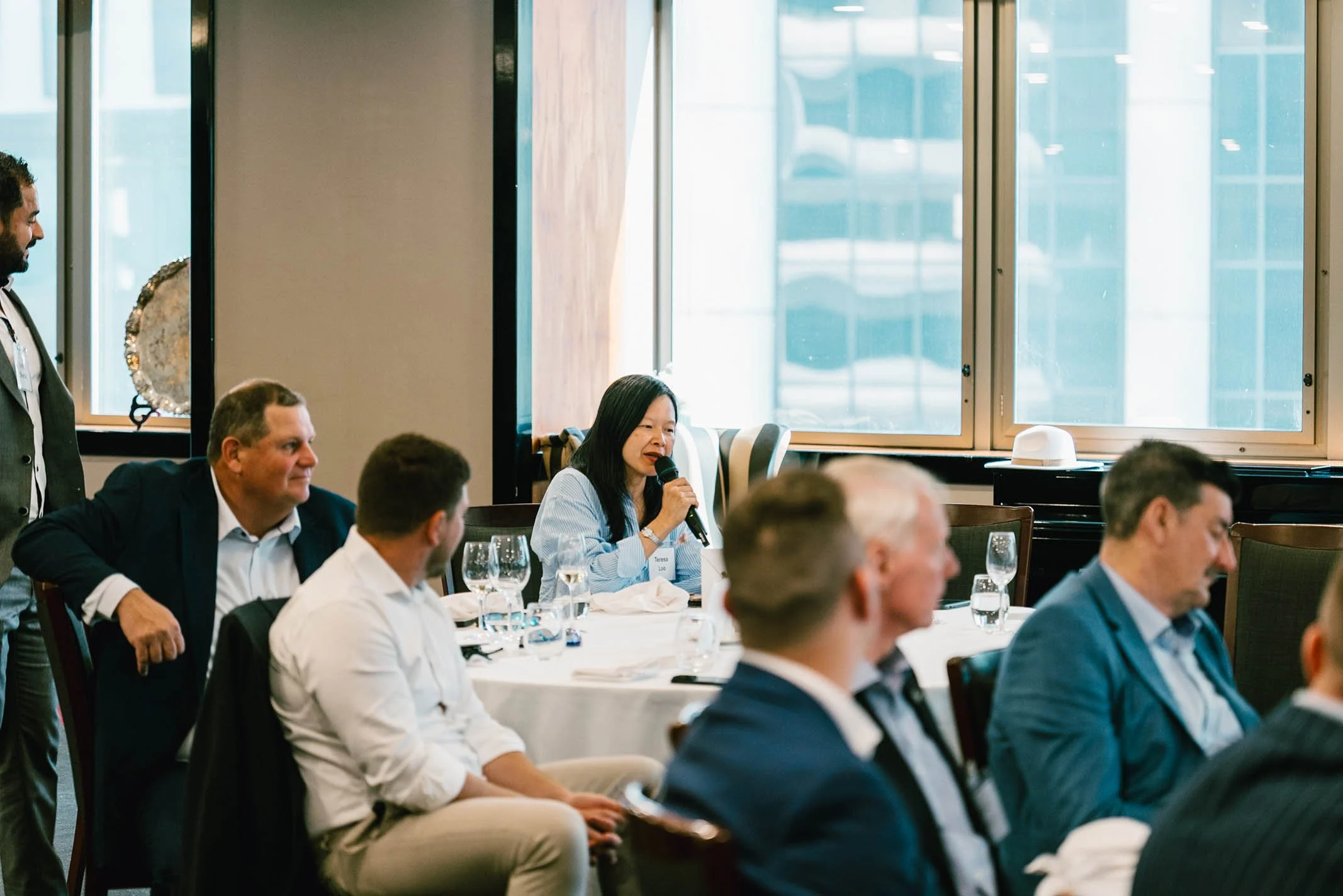 A woman speaking into a microphone at a professional event or conference, surrounded by seated attendees.