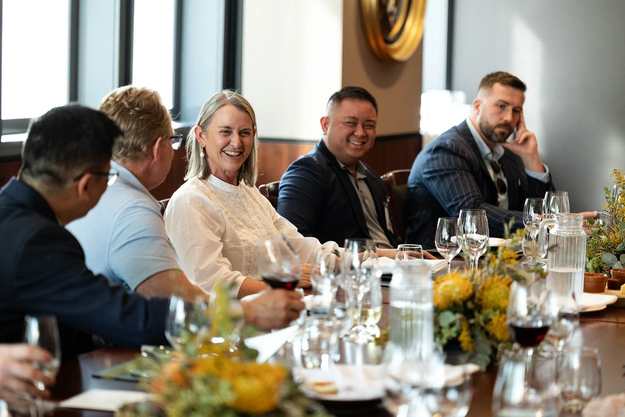 People sitting at a long dining table during a social gathering or meeting, with glasses of wine and floral centerpieces.