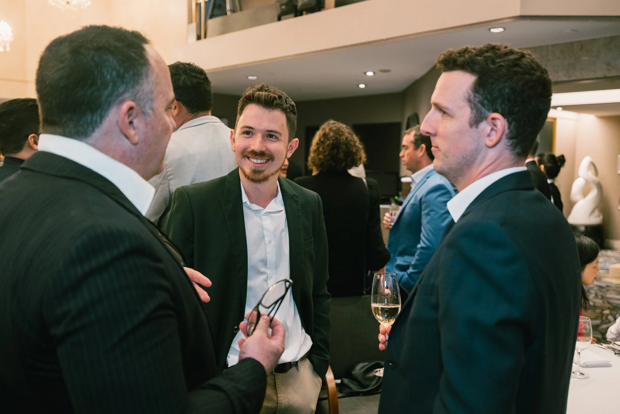 Three men at a formal networking event engaging in conversation, two holding glasses of champagne.