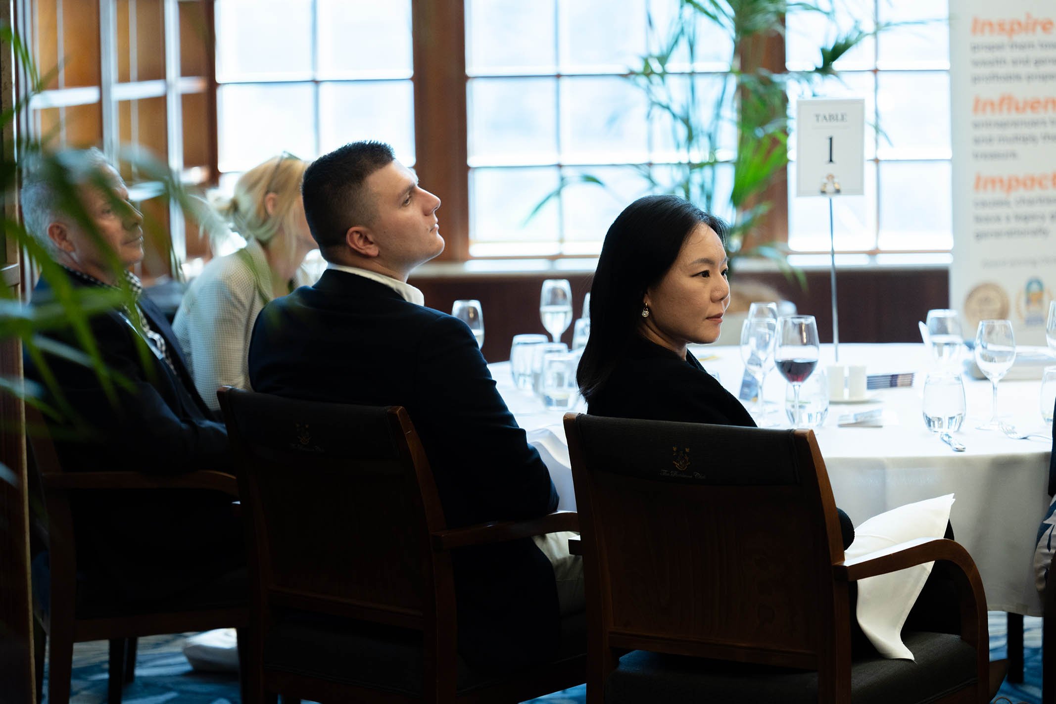 People sitting at a round table during a formal event, with wine glasses and a table sign indicating table 1, in a bright room with large windows and green plants.