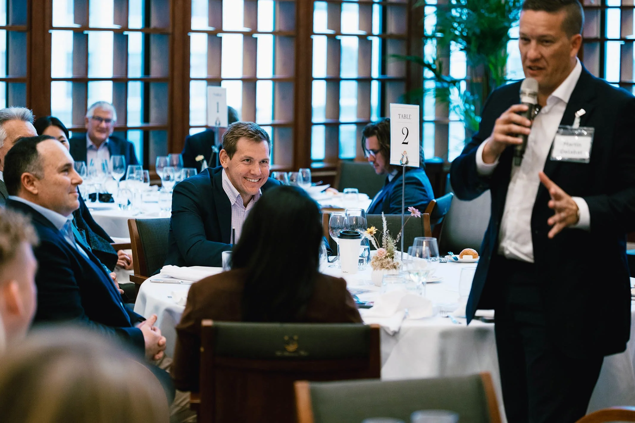 A man in a black suit and white shirt holding a microphone appears to be giving a speech or presentation at a formal event. He is standing near a round table where several people are seated, smiling and listening. The background shows a well-lit room