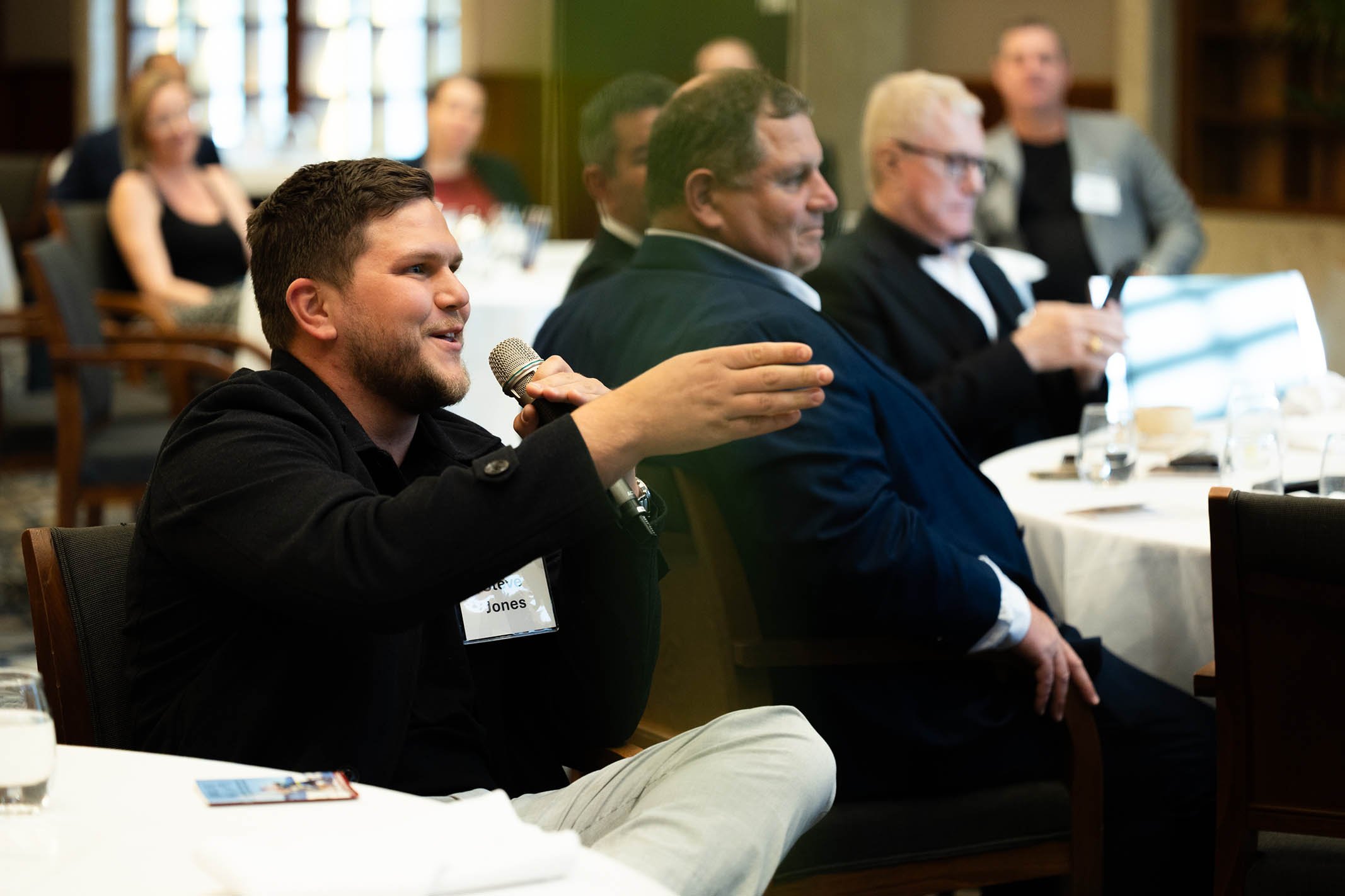 A man with a beard and dark hair holding a microphone speaking at a formal gathering or conference, seated at a table with other attentive men and women in the background.