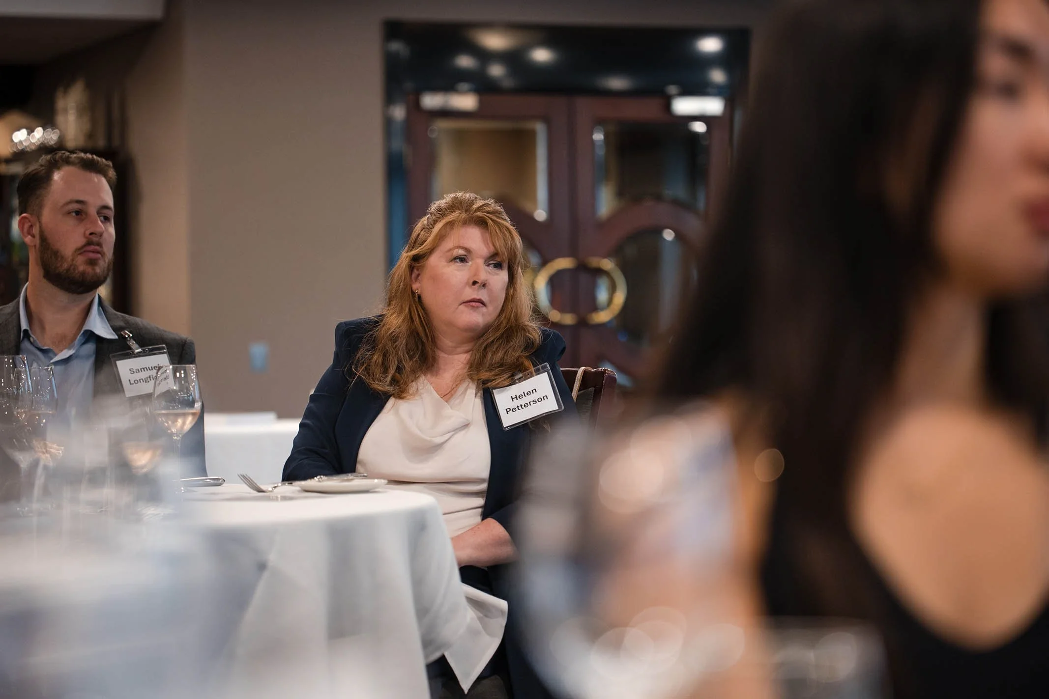 A woman with red hair, wearing a dark blazer and white top, sitting at a round table during a formal event, with a name tag that reads Helen Petterson, in a dimly lit room with other attendees.