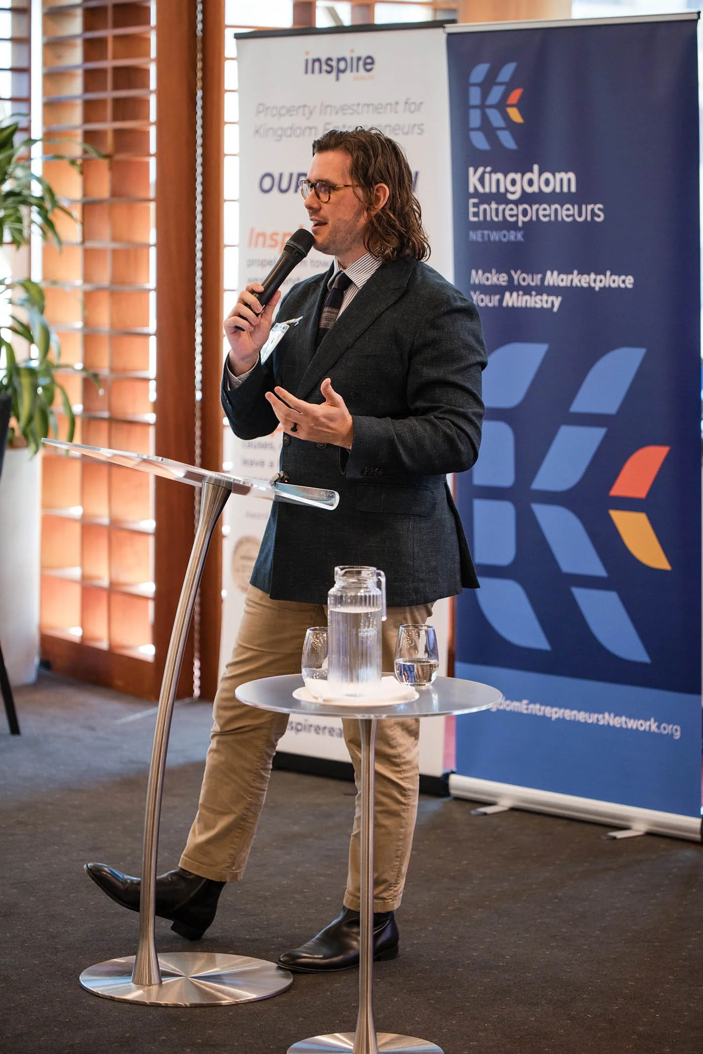 A man in a dark blazer, striped shirt, and beige pants speaking into a microphone at a speaker's podium during a conference, with banners in the background promoting Kingdom Entrepreneurs Network.