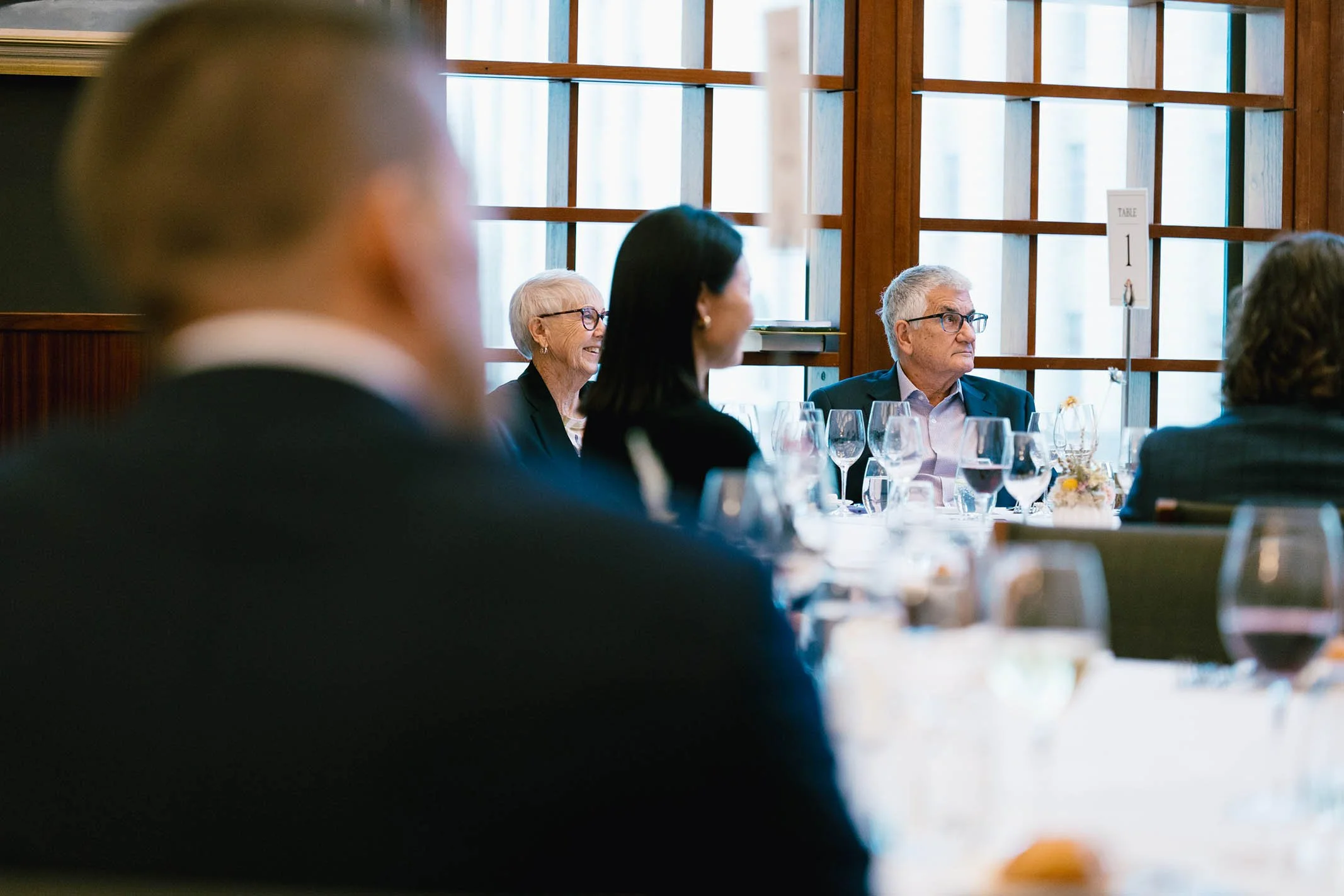 People sitting at a dining table during a formal event, with wine glasses and a table sign visible, in a room with large wooden windowpanes.