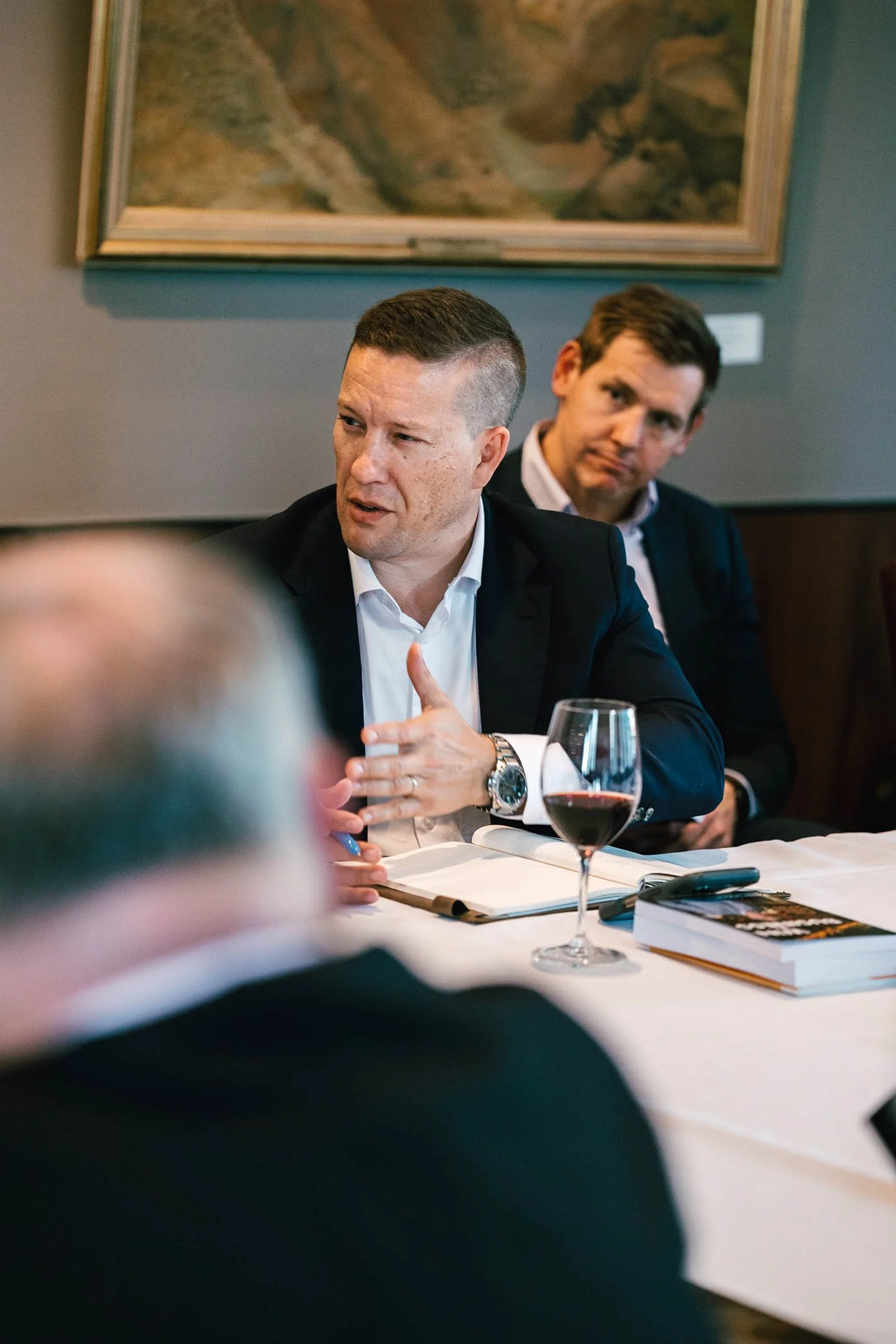Two men in suits sit at a table during a meeting or discussion. One is speaking, with open hand gestures. There is a glass of red wine, an open notebook, and a book on the table. The background features a framed painting or picture on the wall.
