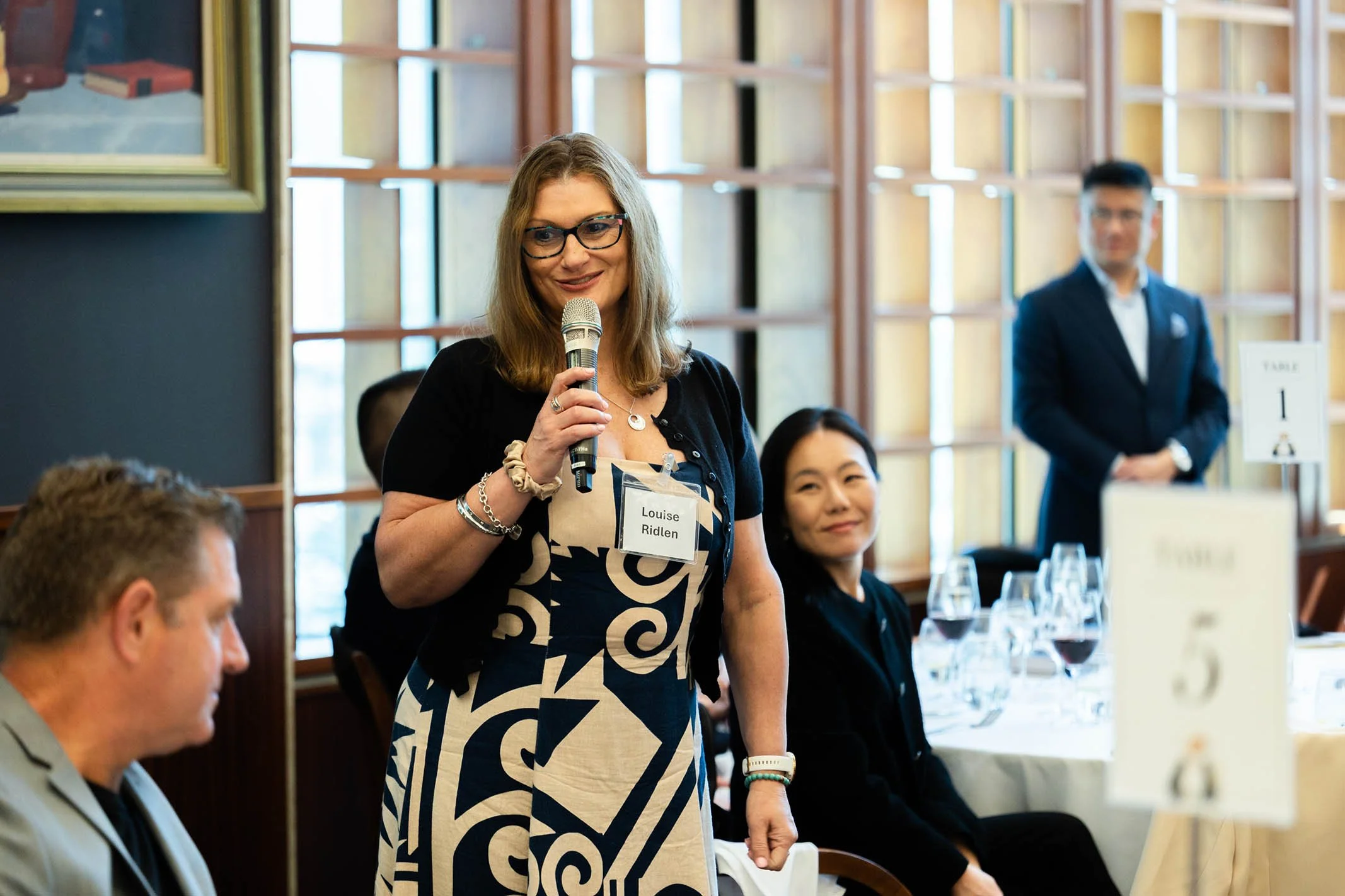 Woman with glasses, wearing a black top and patterned dress, speaking into a microphone at a formal event. People are seated at tables with wine glasses, and there is a man in a suit standing in the background.