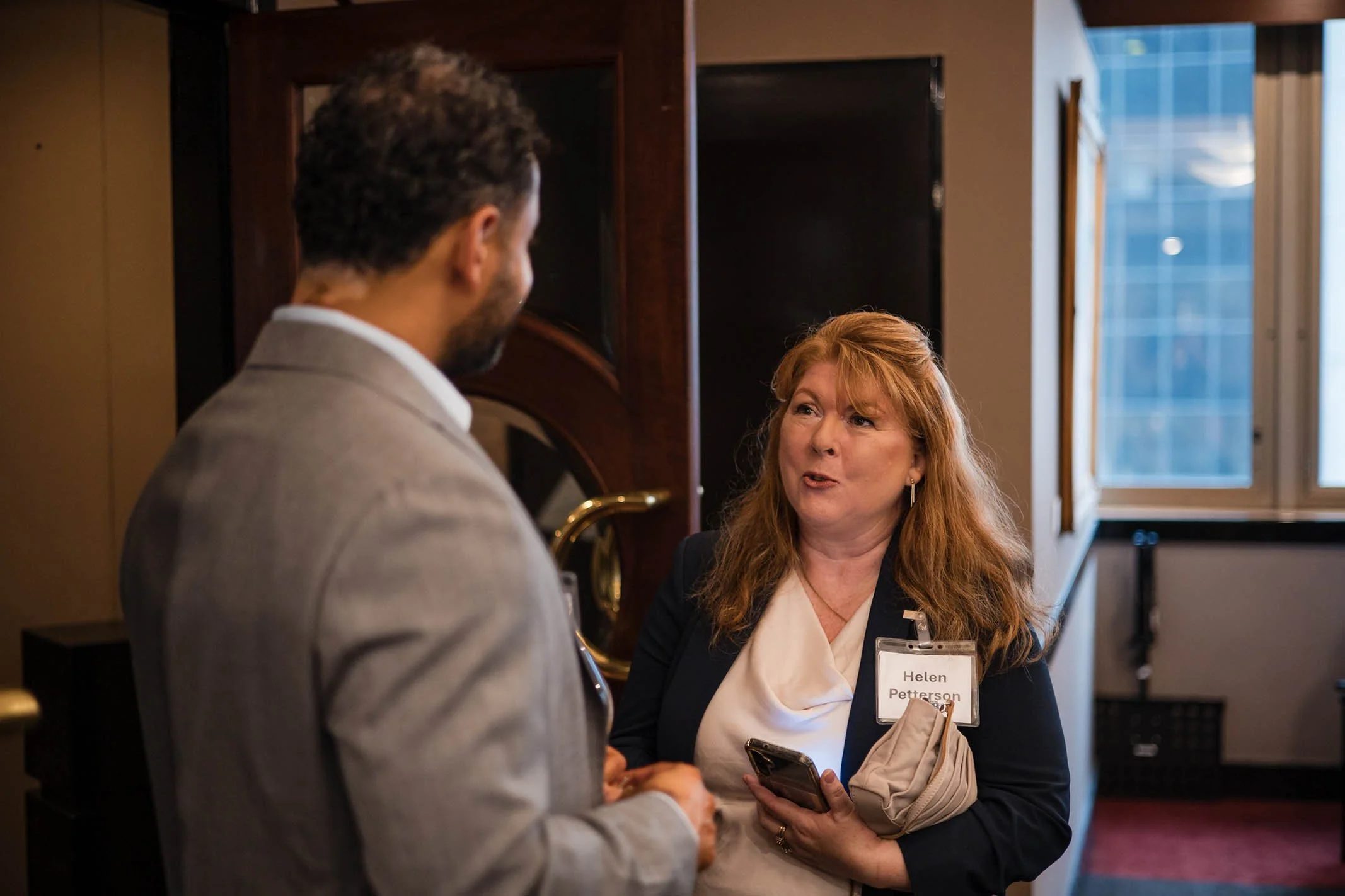 A woman with red hair, wearing a dark blazer and a name tag that reads 'Helen Petterson,' is holding a phone and talking to a man dressed in a gray suit. They are indoors, in a professional setting near a window.