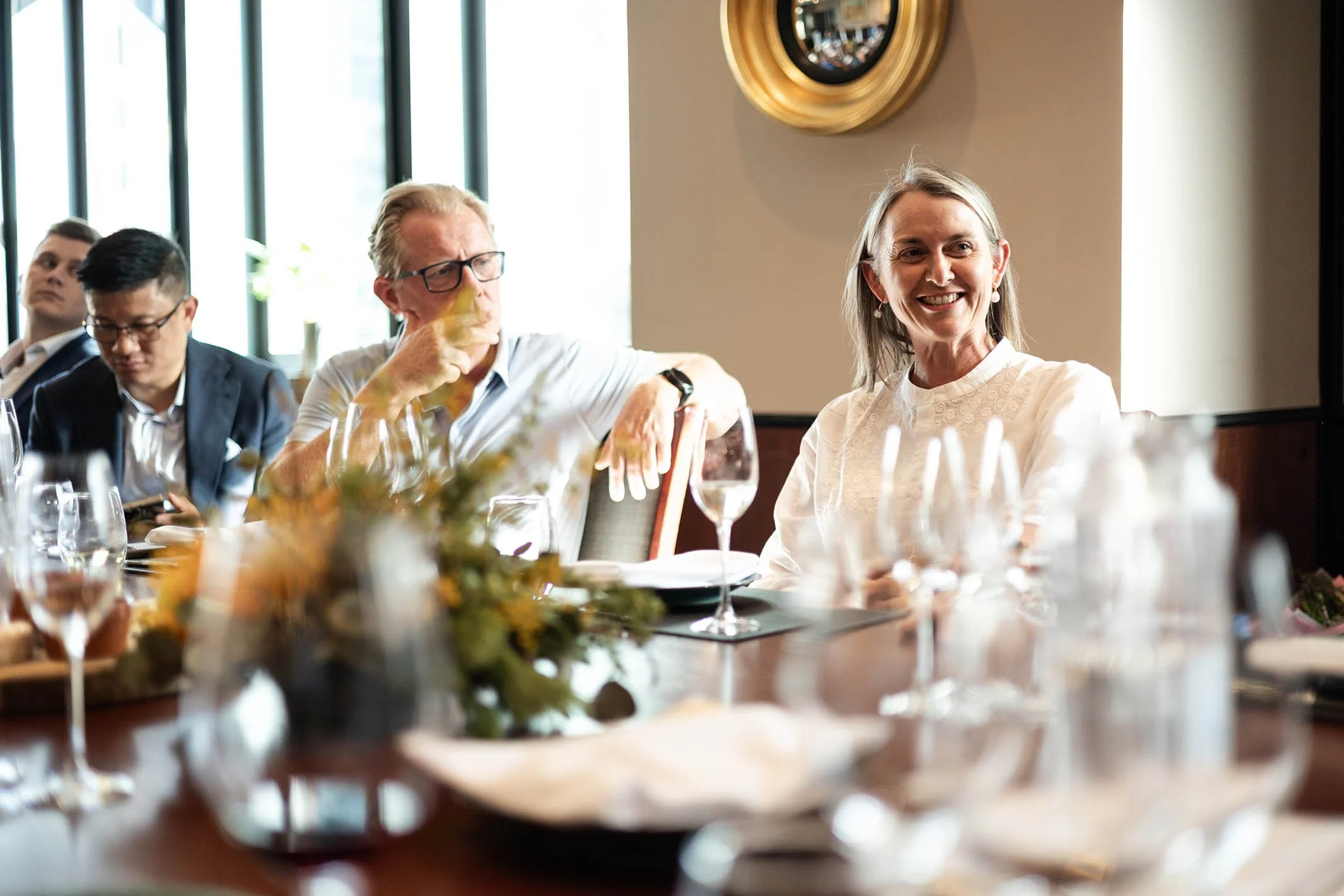 People sitting at a table in a restaurant or banquet hall, with glasses of wine, flowers, and tableware, engaged in conversation and smiling.