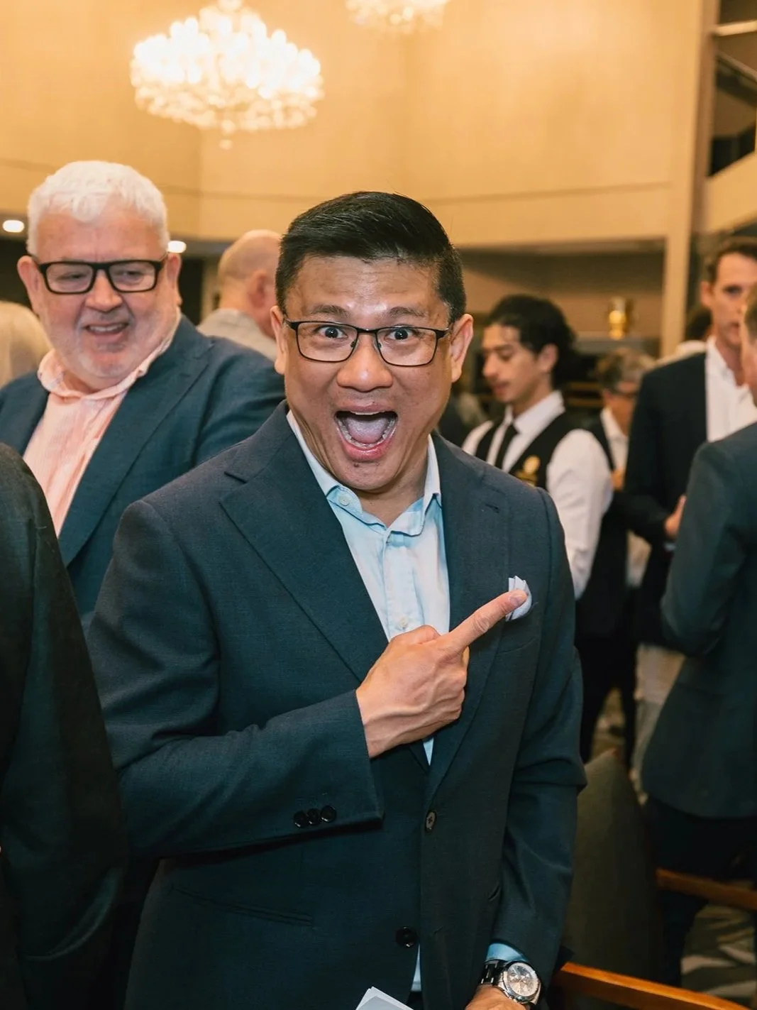 A man with glasses in a suit smiling and pointing to his chest at a formal event with other people in the background.