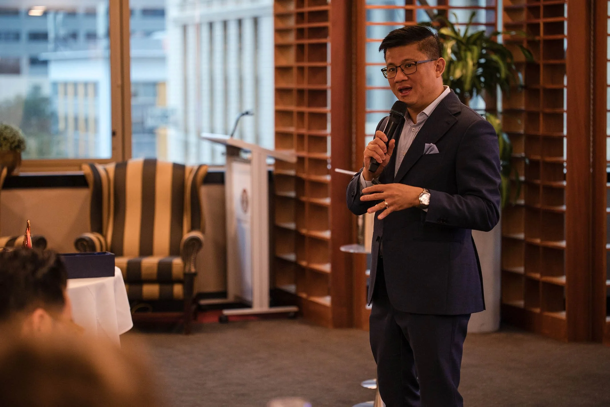 A man in a dark suit with glasses and a watch is speaking into a microphone in a wood-paneled room with windows and plants, addressing an audience.