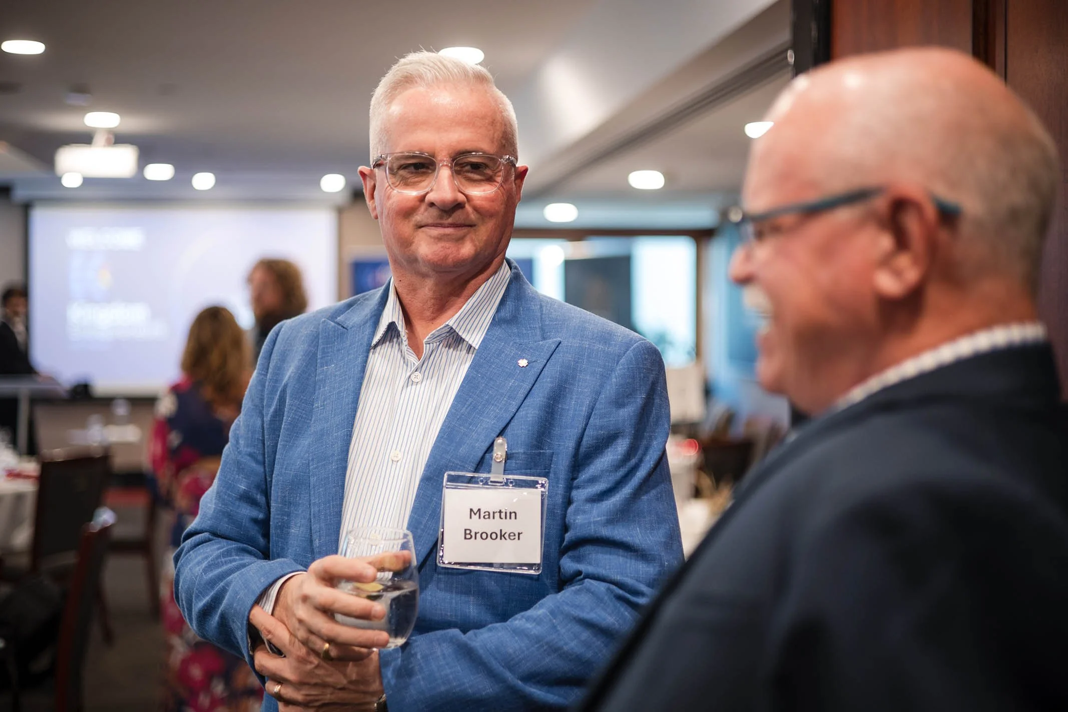 Two men are engaged in conversation at a professional event. The man on the left, wearing a blue blazer and glasses, holds a glass of water and has a name badge that reads 'Martin Brooker.' The man on the right, with a blurred face, is listening and 