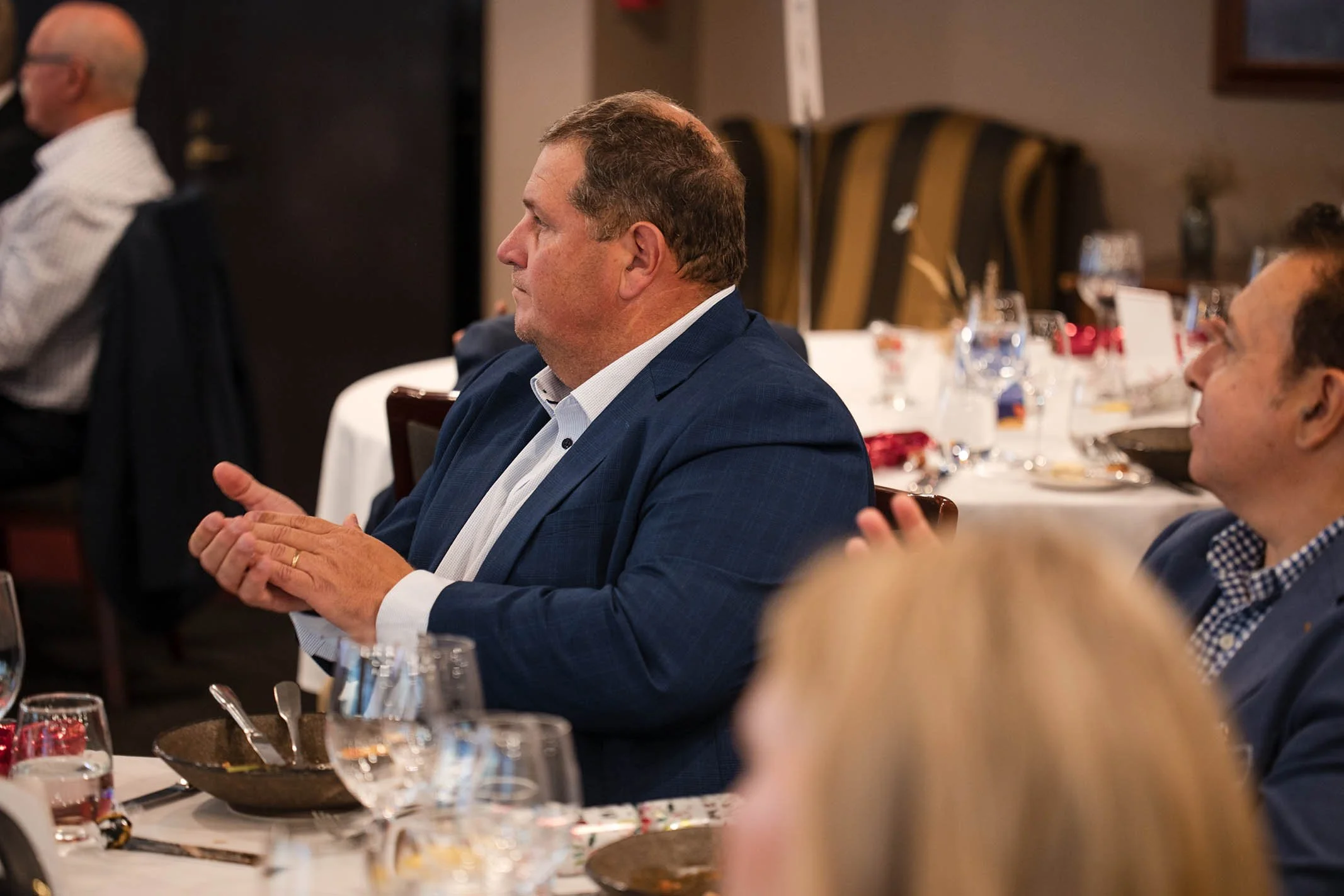 A man in a blue suit and white shirt sitting at a dinner event, clapping his hands.