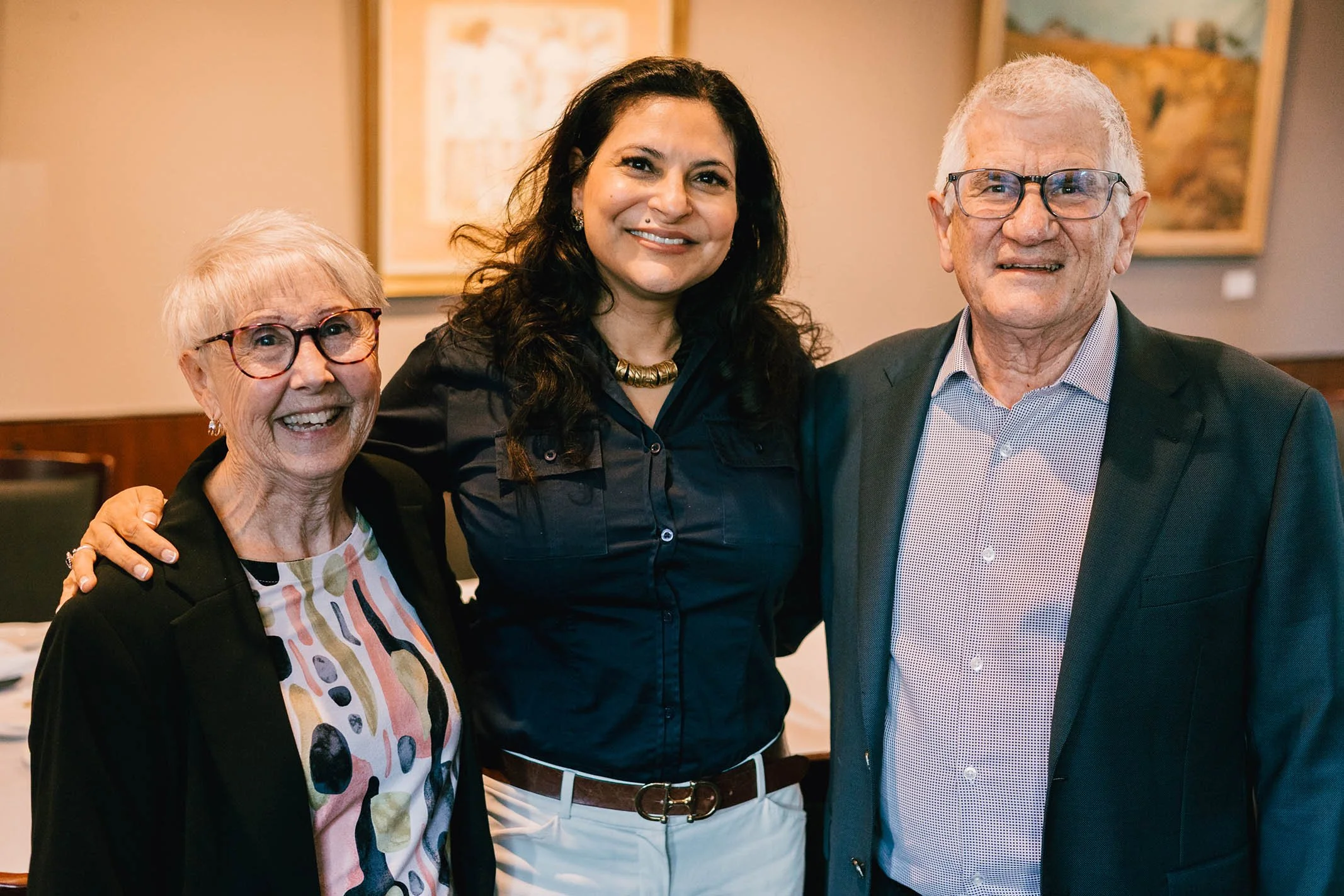 Three smiling people, two women and one man, standing close together indoors with artwork on the wall behind them.