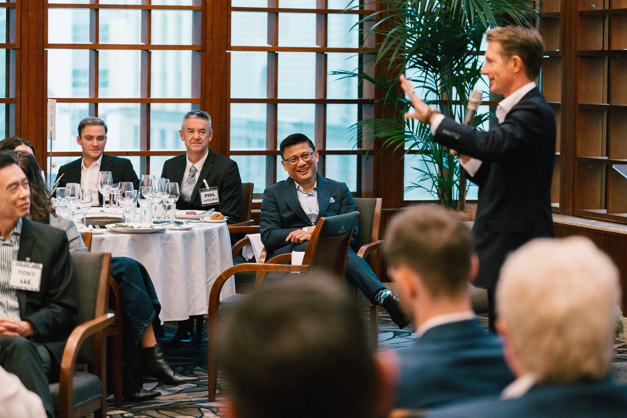 A man in a black suit giving a presentation with a microphone in a conference room with seated attendees listening and smiling.