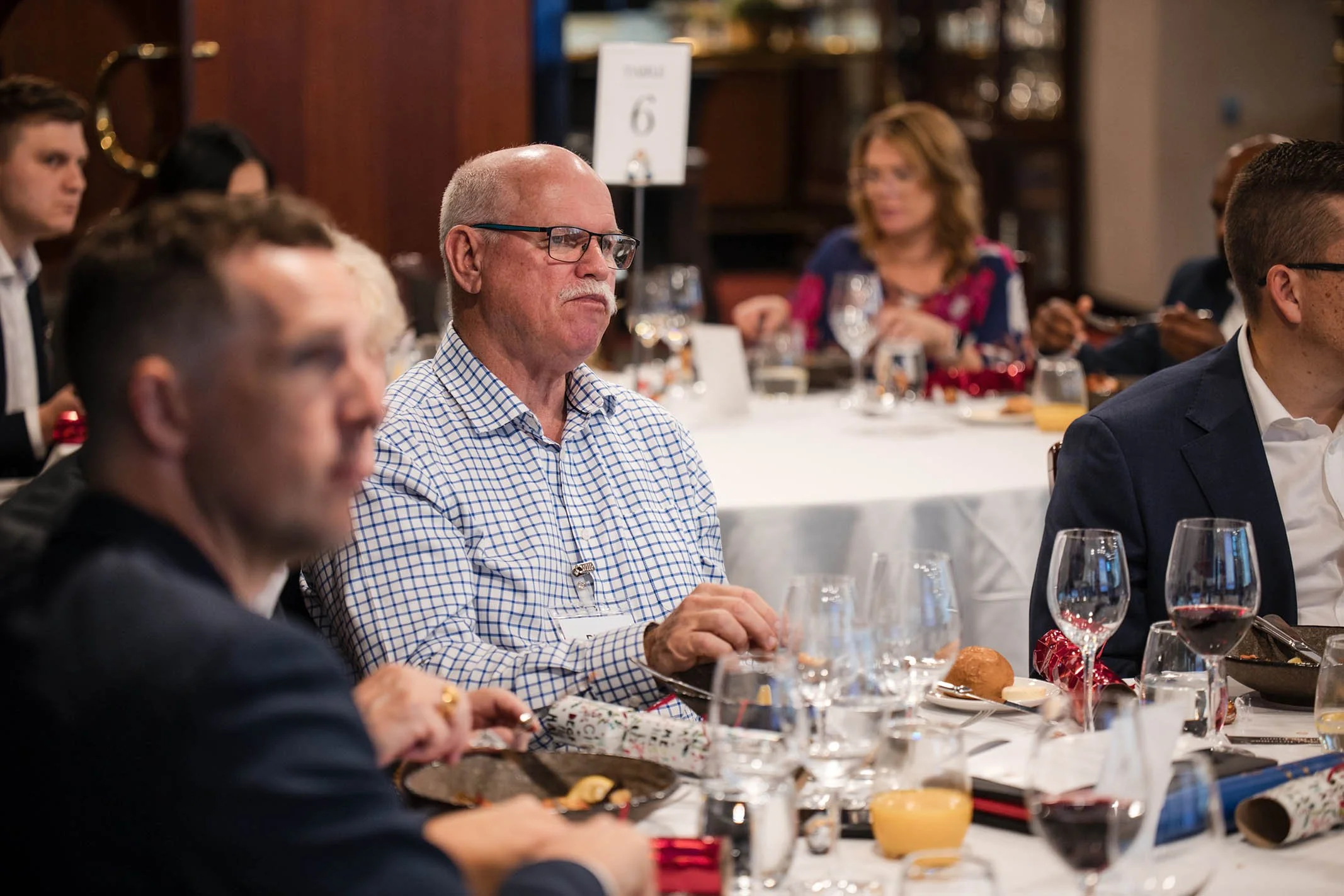 A group of people sitting around a banquet table at a formal event, with food and drinks, some with glasses of red wine, in a decorated room.