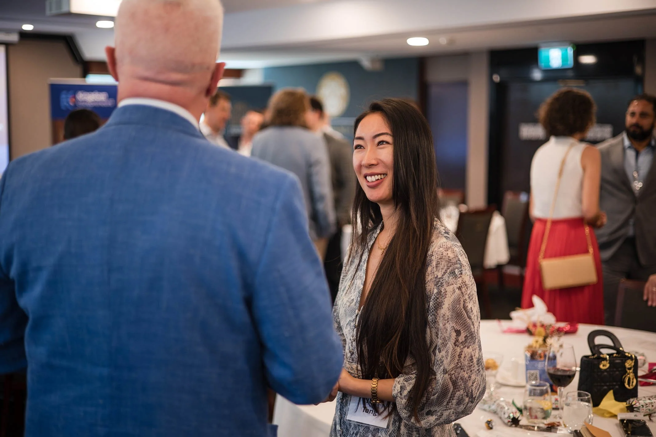 A woman with long dark hair smiling and talking to a bald man dressed in a blue suit at a social gathering or conference.