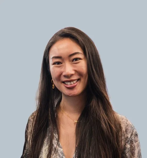 A woman with long dark hair smiling at the camera, wearing a patterned blouse and gold earrings, against a plain light blue background.