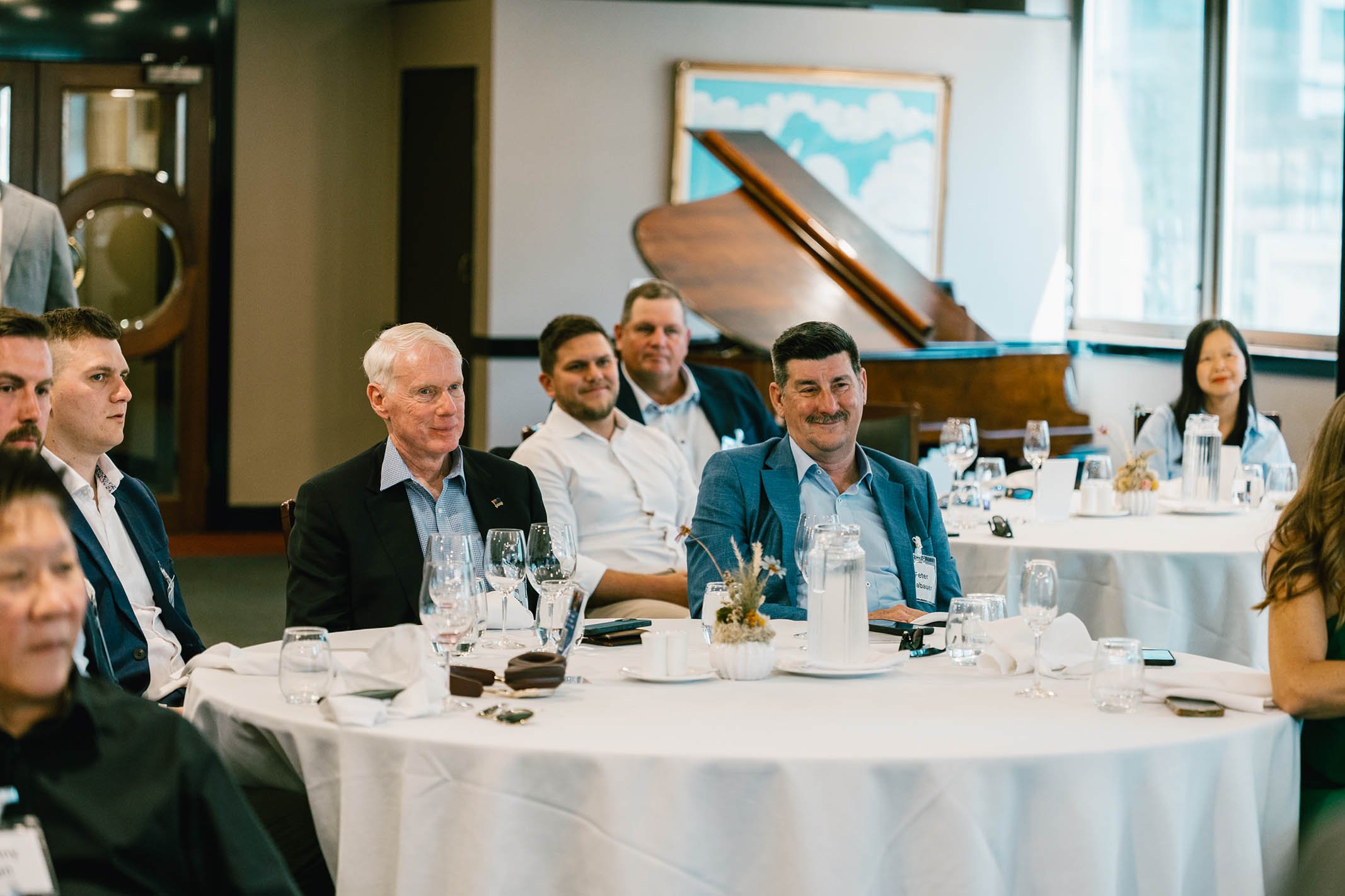 People attending a formal event or meeting sitting around round tables with white tablecloths, glasses, and floral centerpieces, in a room with large windows and a grand piano in the background.